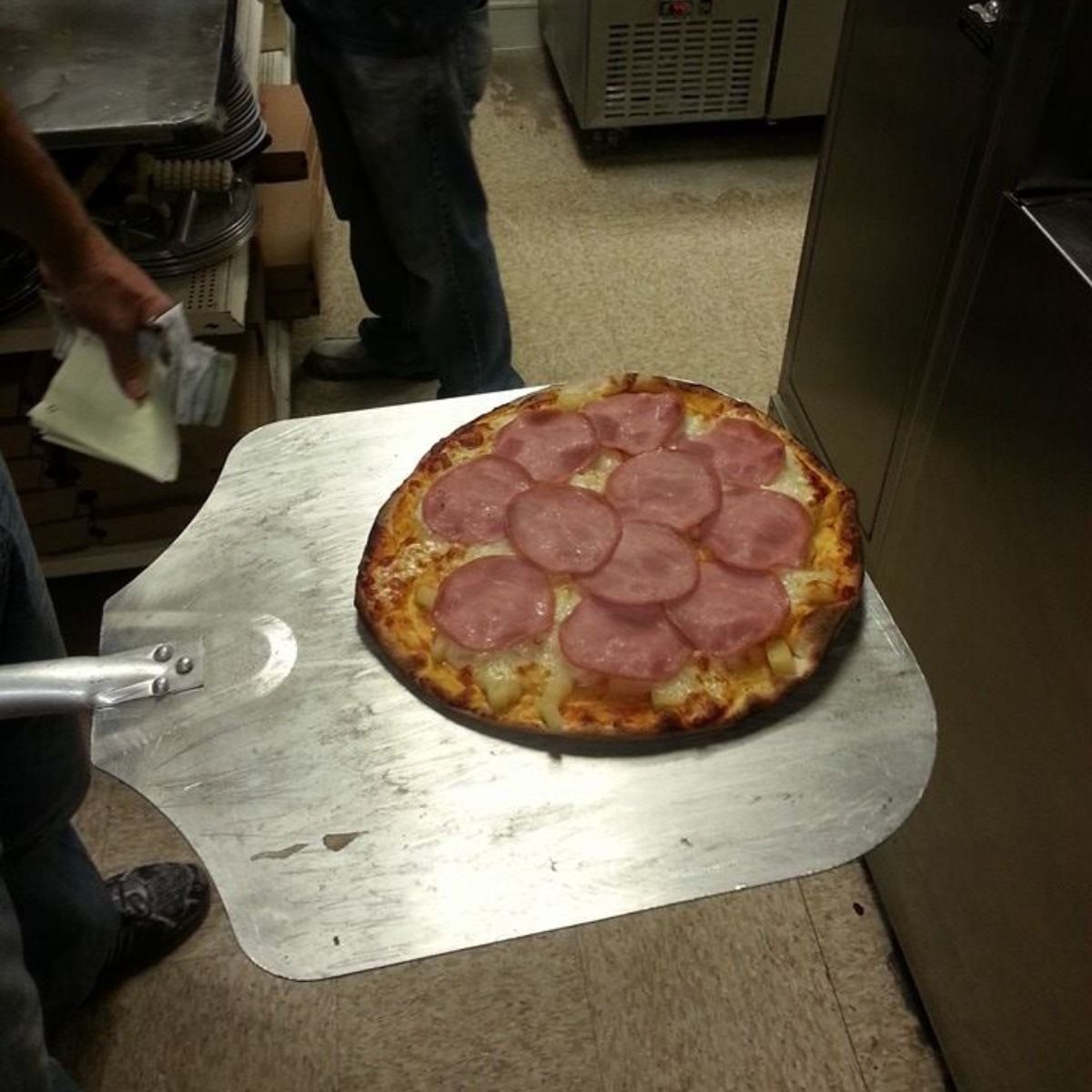 Pizza with ham on a metal peel, person holding it, other person in background near kitchen.