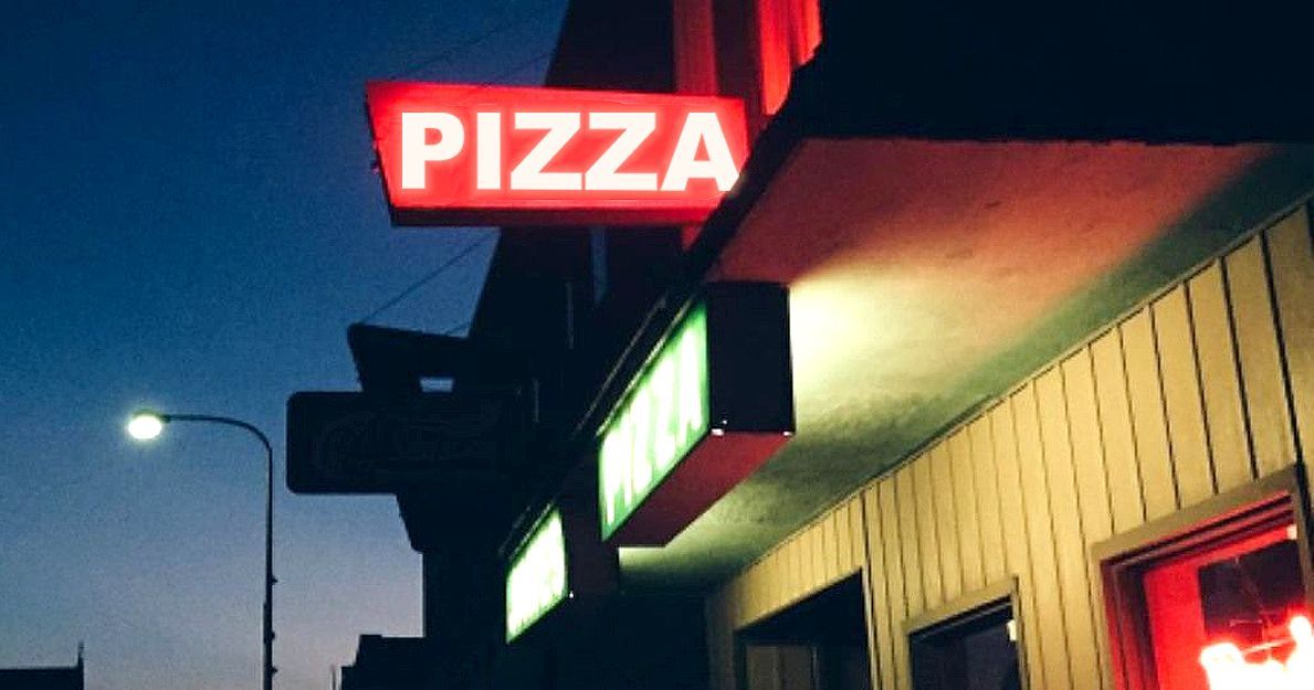 Neon "PIZZA" sign on a building at dusk; streetlights and dark blue sky.