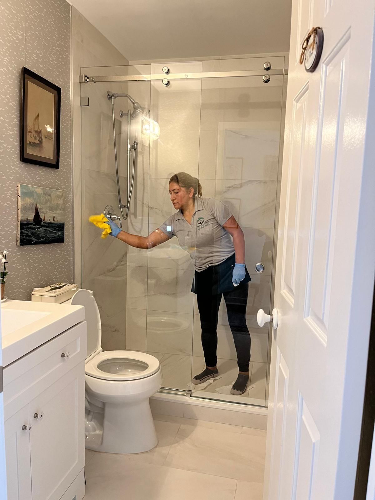 A person wearing gloves cleaning a shower. Bathroom with toilet and sink in view.
