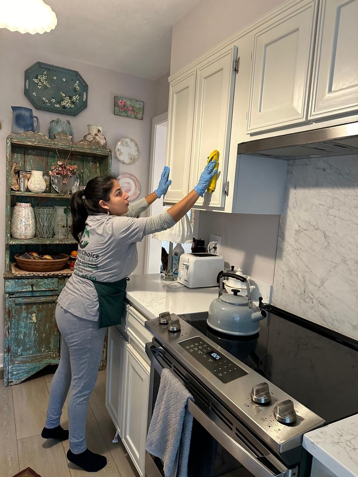 Woman cleaning kitchen cabinets, wearing gloves. Grey sweatsuit. Bright kitchen.