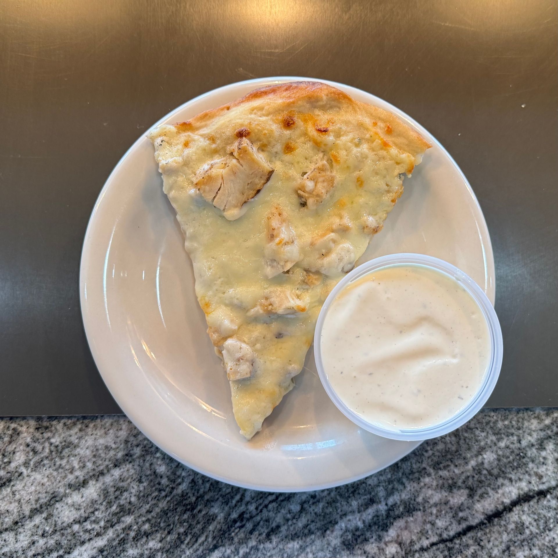 Slice of pizza on a white plate with a small cup of white dipping sauce on a countertop