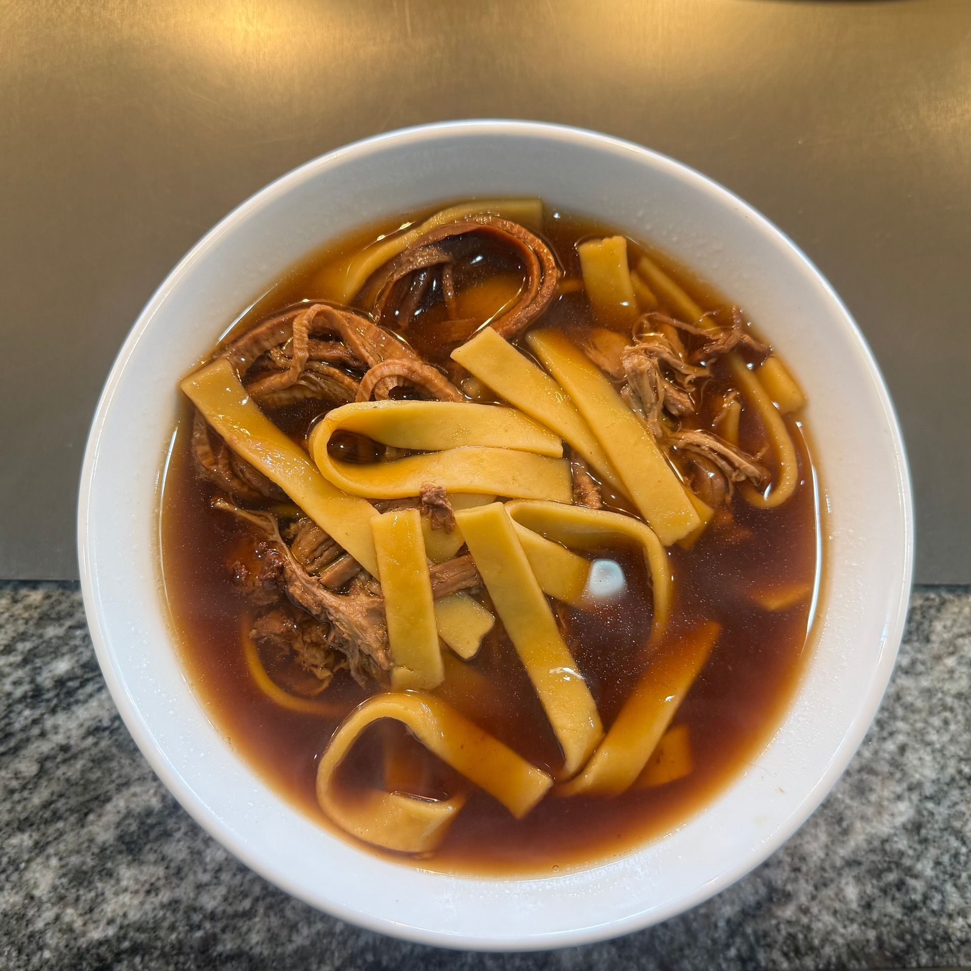 Bowl of dark broth noodle soup with wide noodles on a table