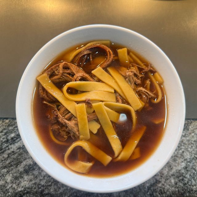 Bowl of brown noodle soup with wide flat noodles on a table.