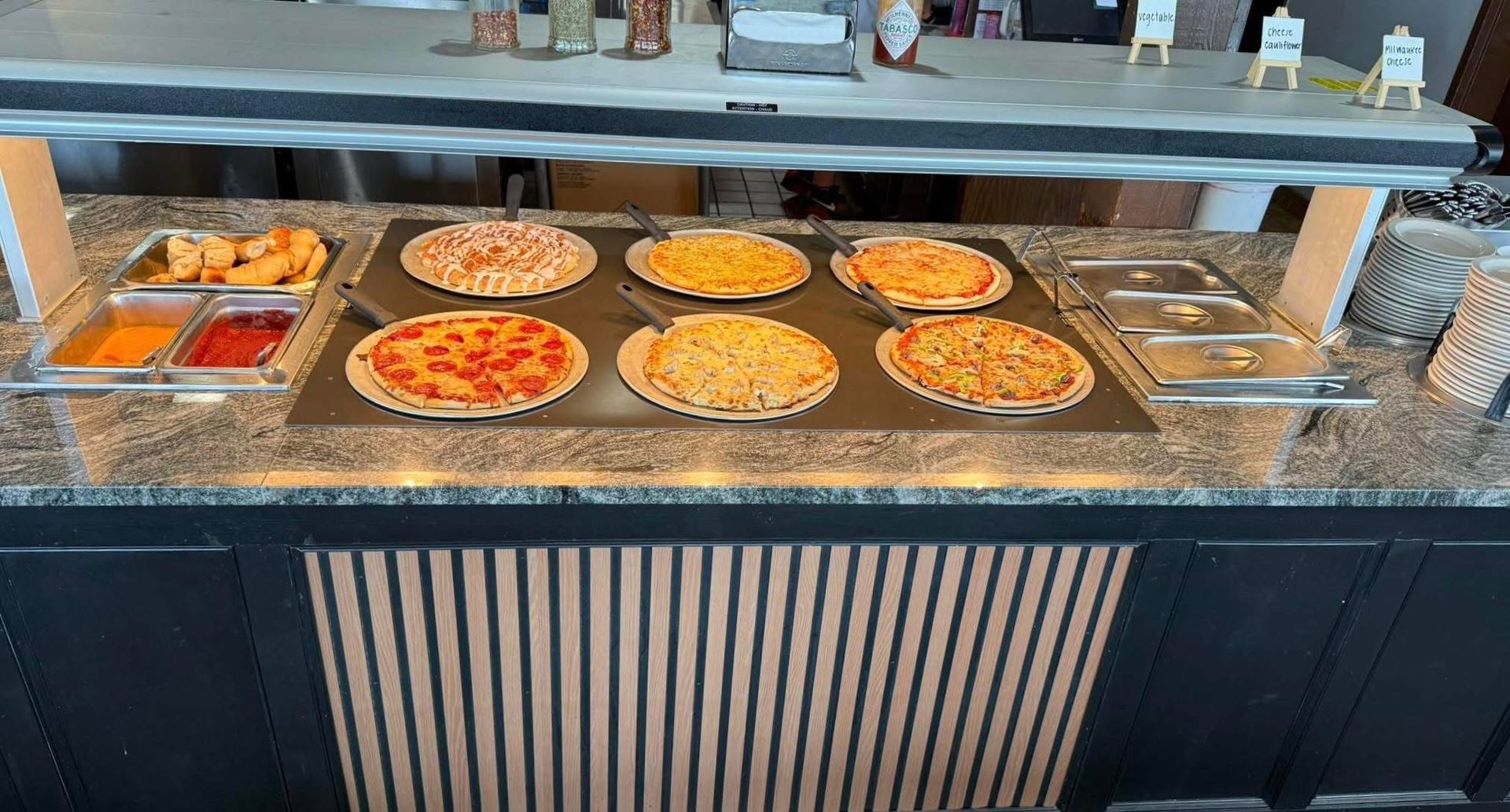 Buffet counter with trays of pizza and a small dessert tray behind glass at a restaurant.