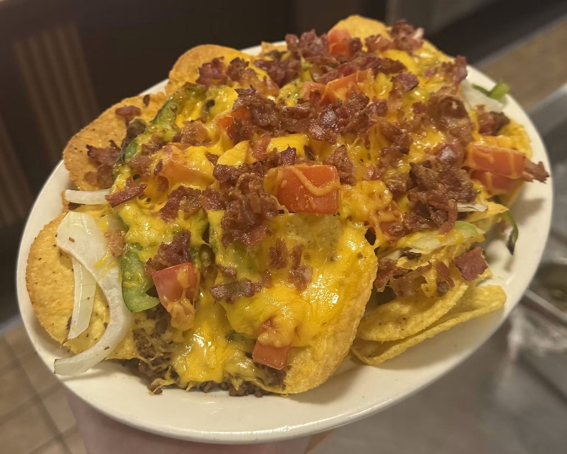 Loaded nachos topped with melted cheese, bacon, tomatoes, and green onions on a white plate.