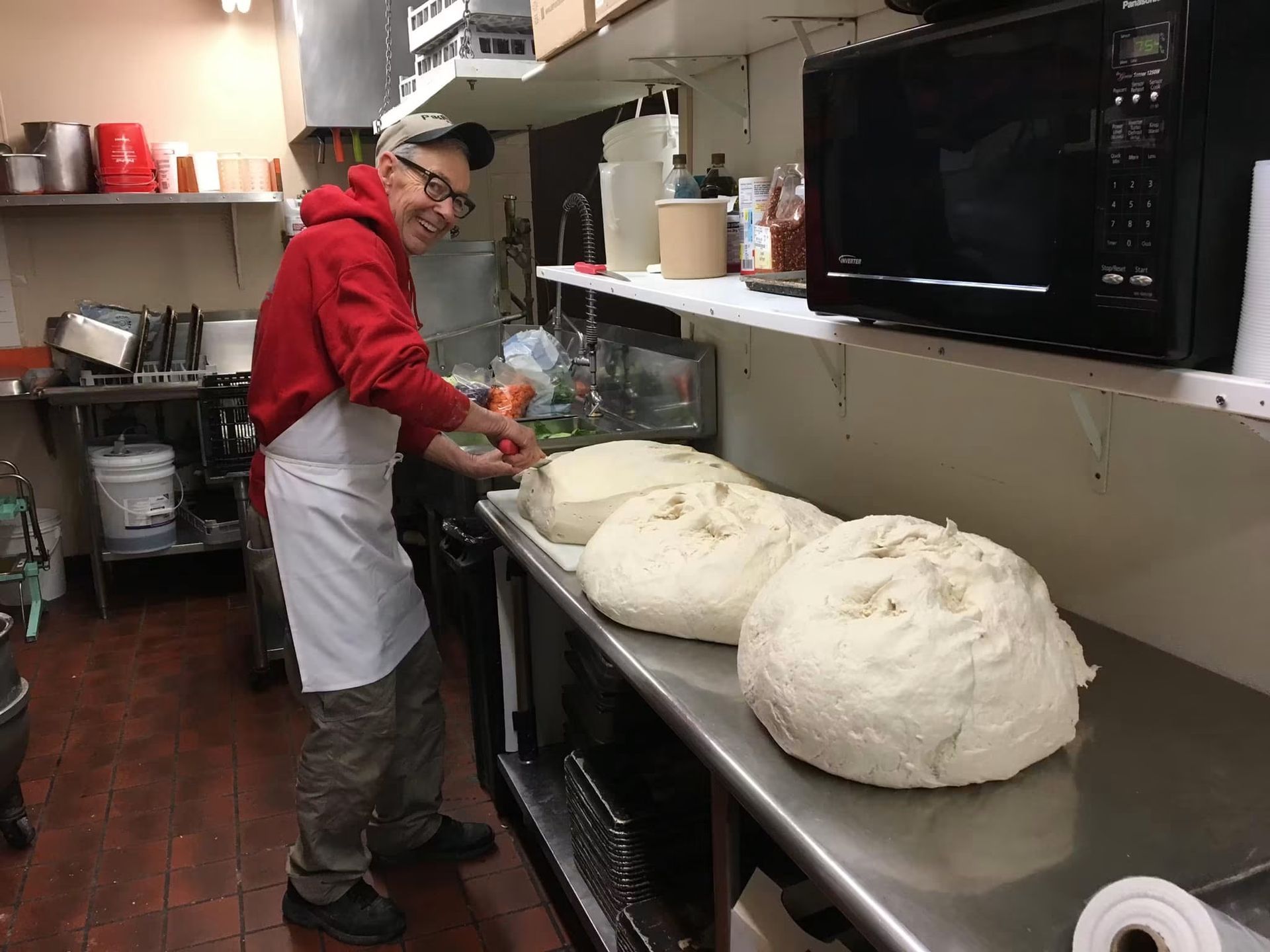 Person in a red hoodie and apron works in a kitchen beside large dough balls on a counter.