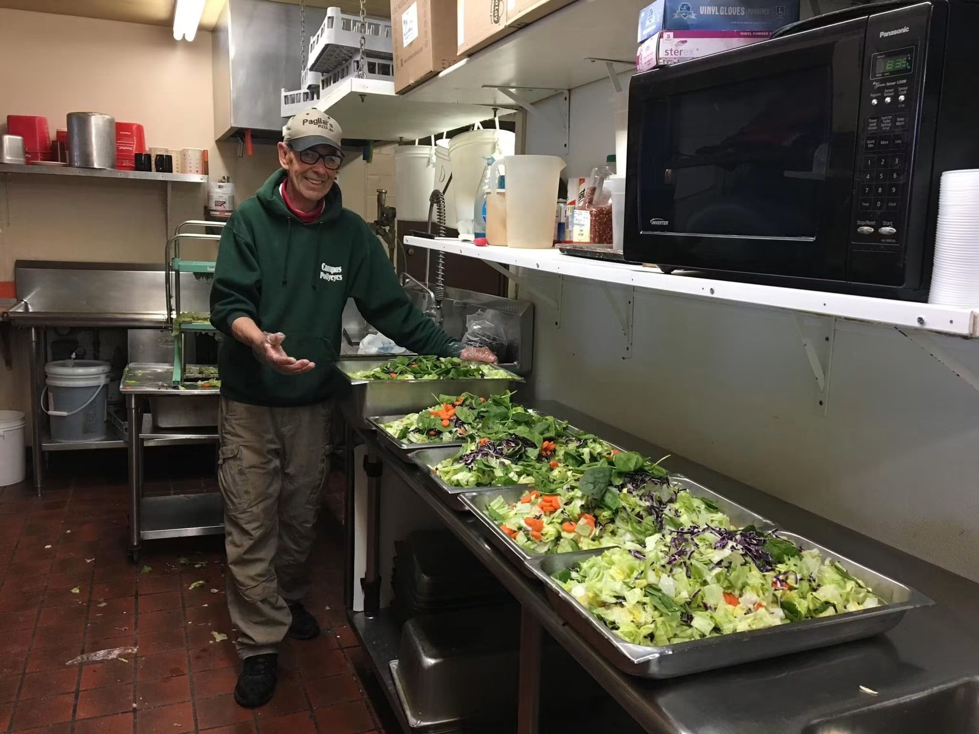 Person in a kitchen holding a tray of salad ingredients beside a counter of prepared salads