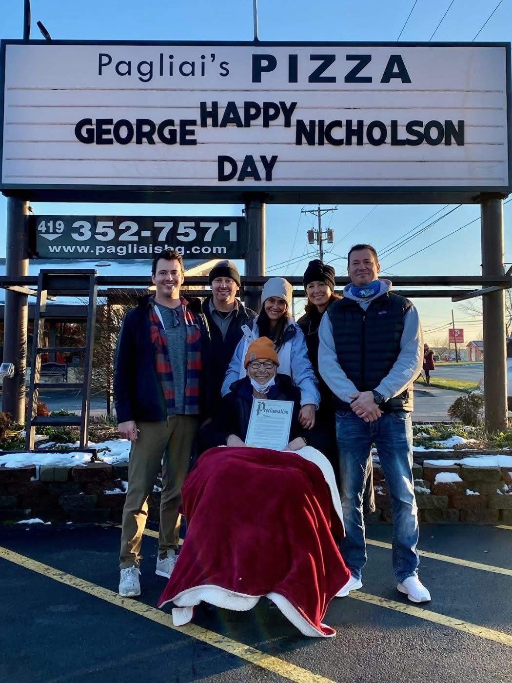 Four people pose outside Pagliai’s Pizza sign in winter, with a red-draped object in front.