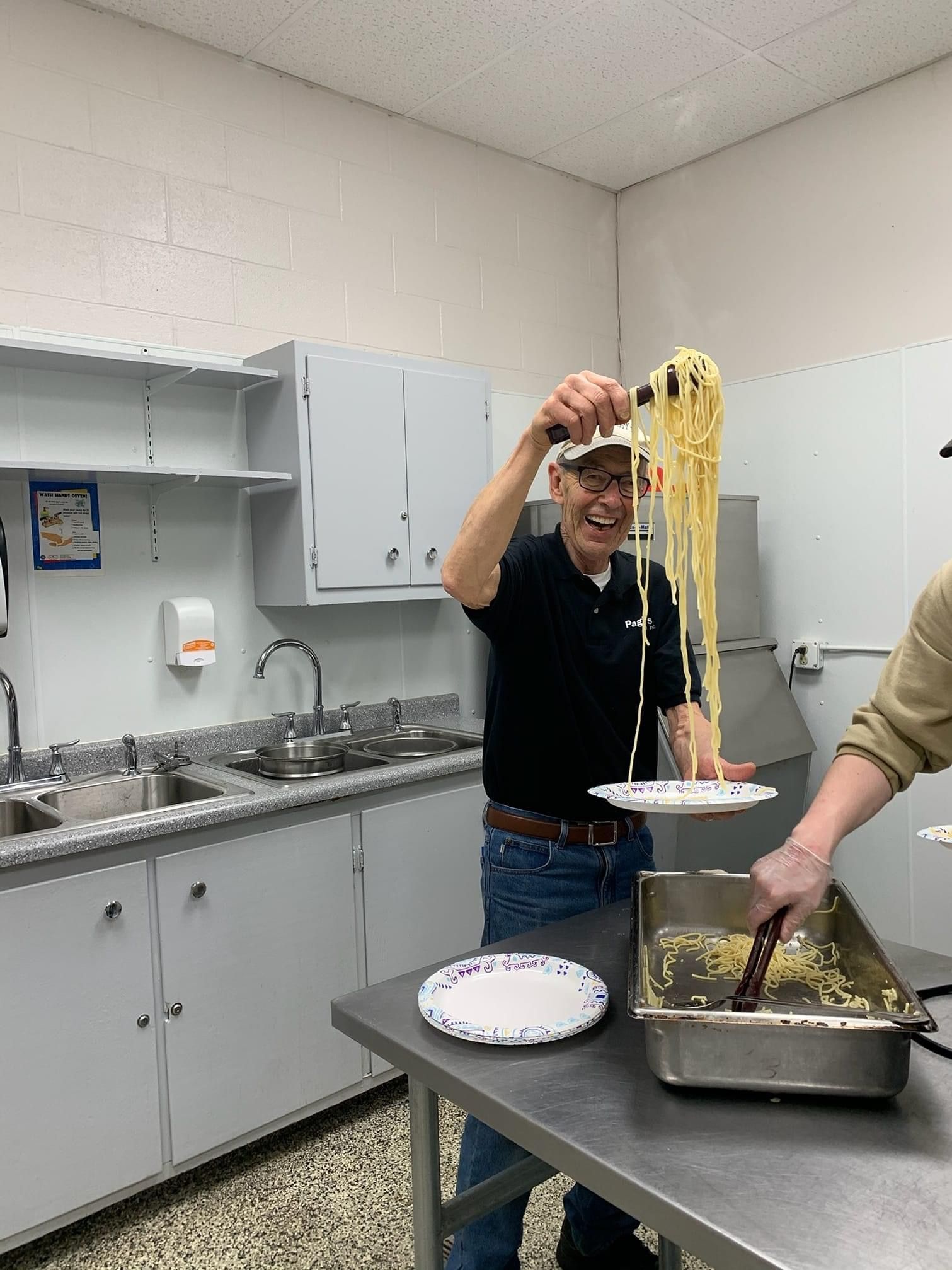 Two people serve noodles in a kitchen, with a tray and plates on the counter.