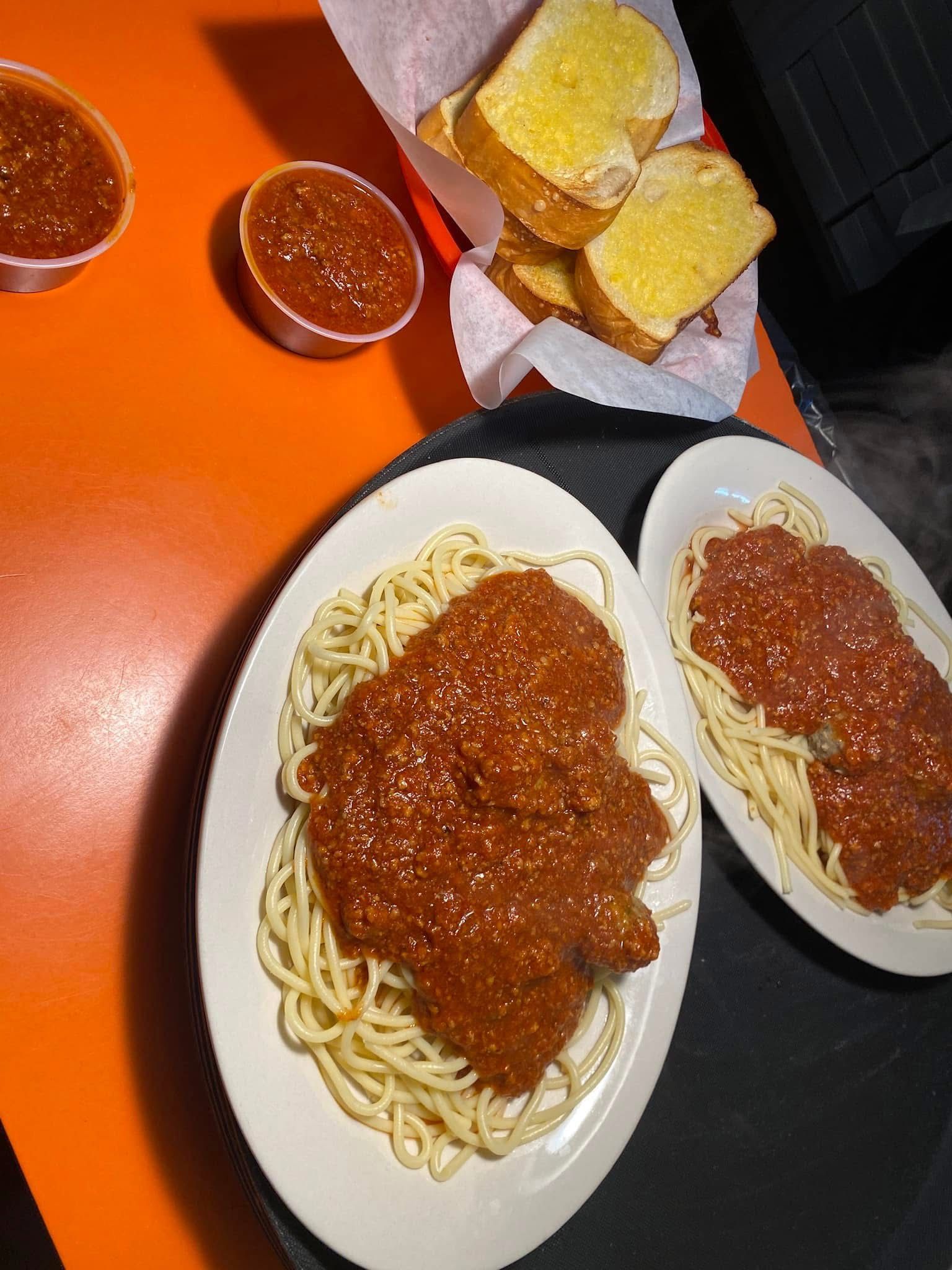 Two plates of spaghetti with meat sauce and bread on an orange table, with cups of sauce nearby