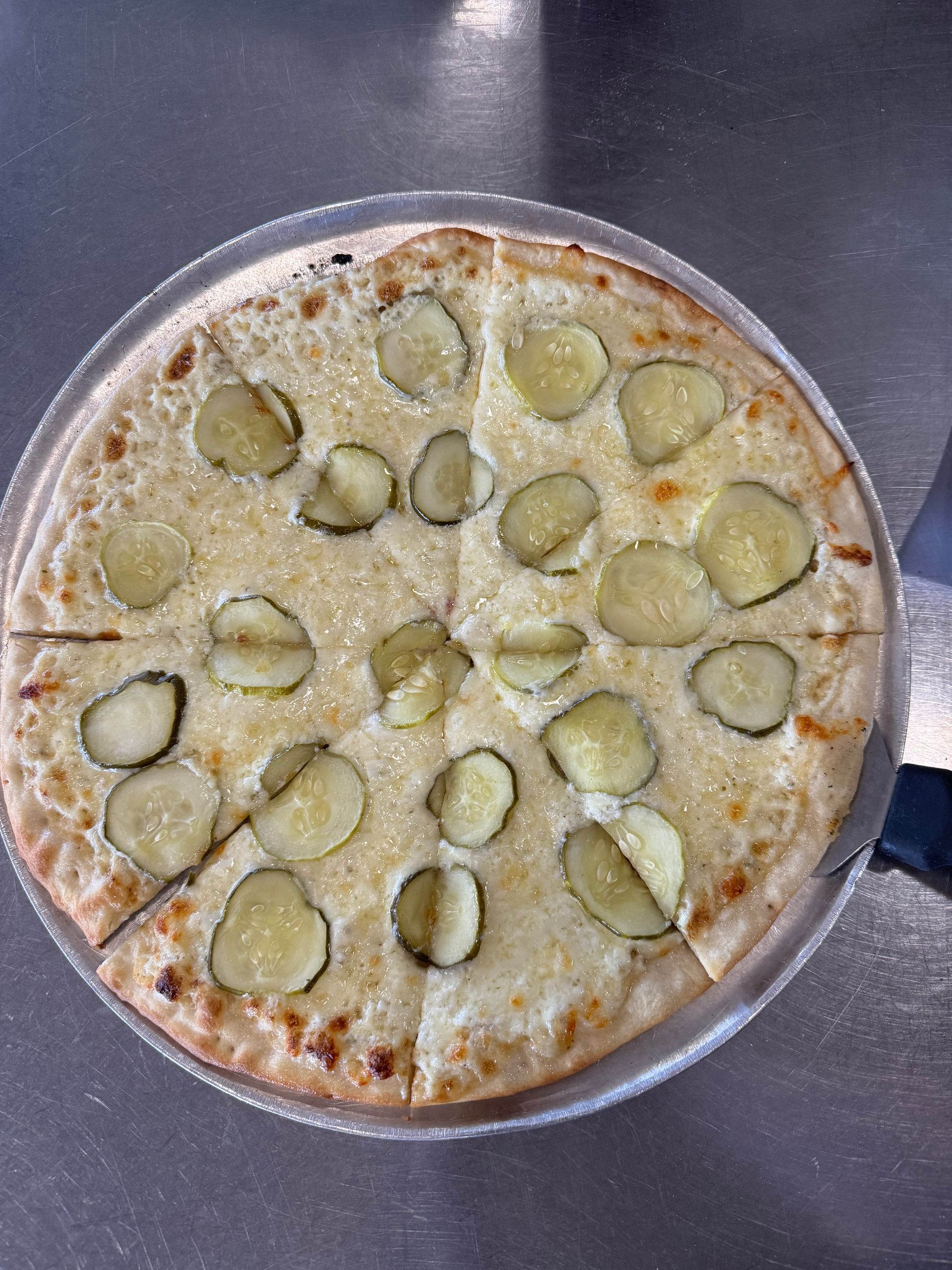 Round cheese pizza topped with sliced pickles on a metal pan against a dark background