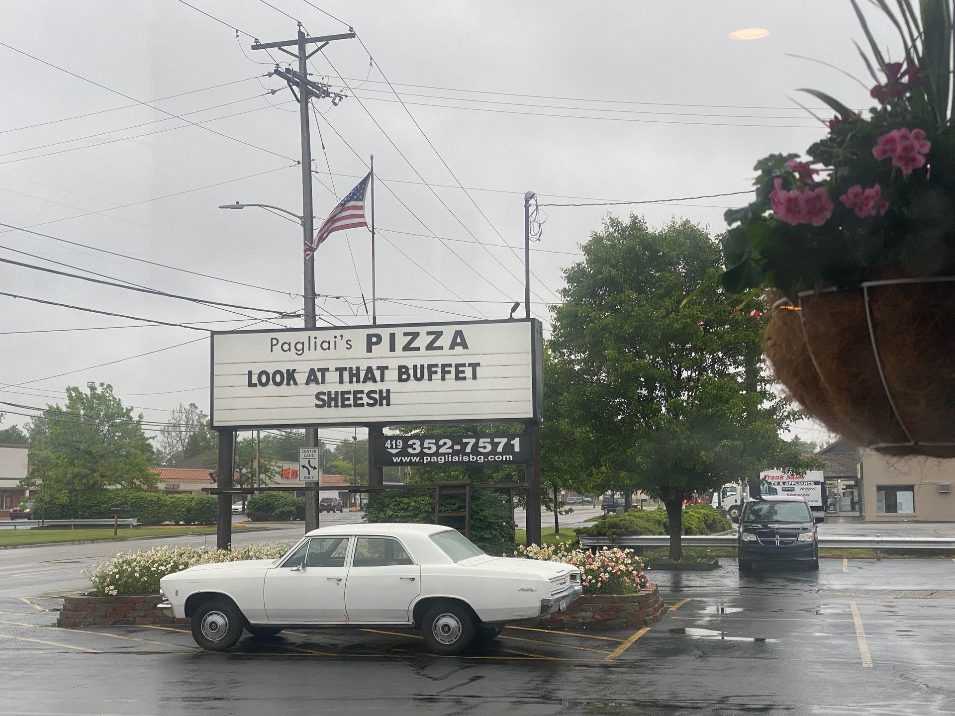 White car in rainy parking lot beside a pizza buffet sign and hanging flower basket.