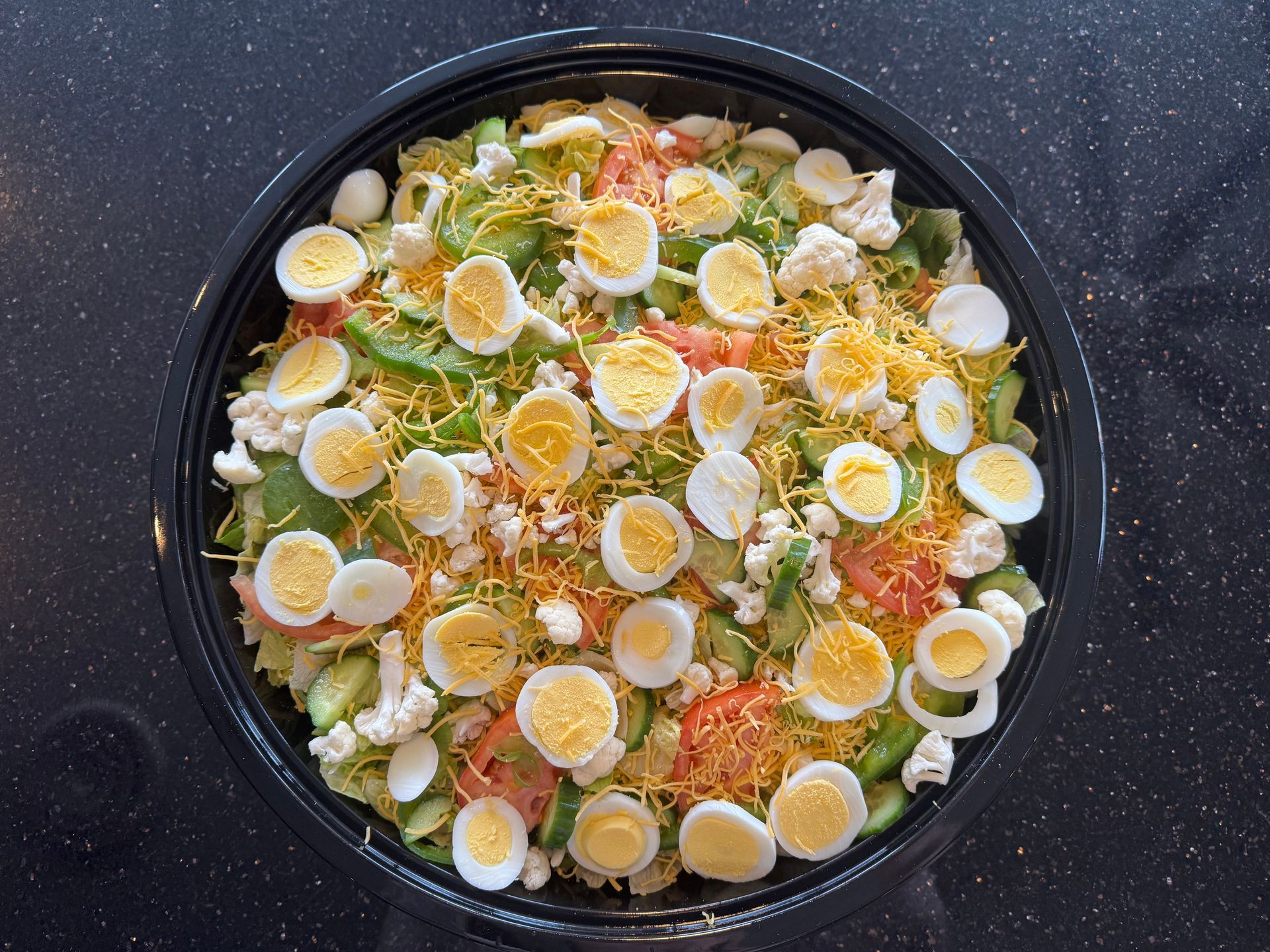 Salad in a black bowl topped with sliced boiled eggs and shredded cheese on a dark surface