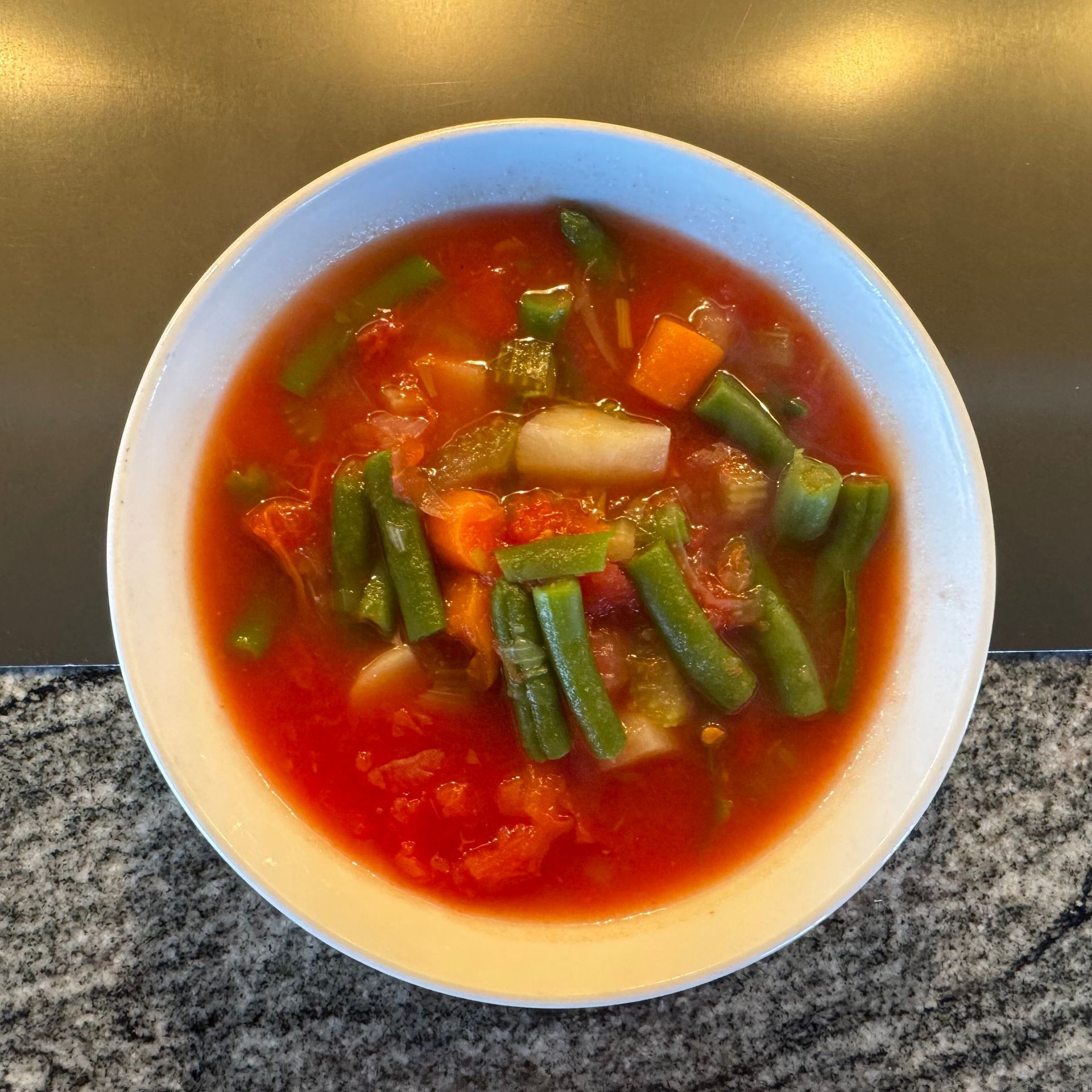 Bowl of red vegetable soup with green beans and diced vegetables on a gray countertop