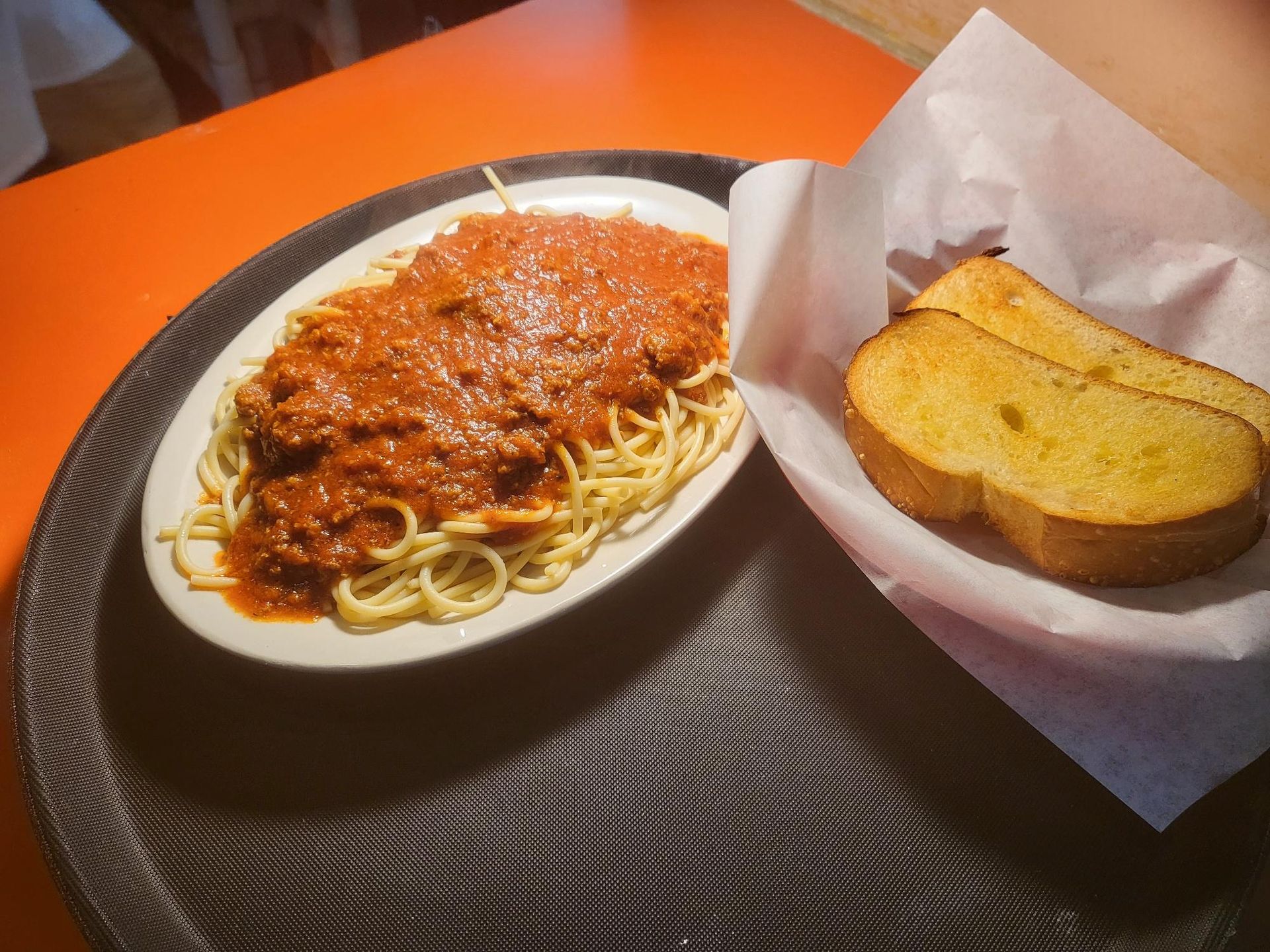 Spaghetti with red sauce and garlic bread on a black tray atop an orange table.
