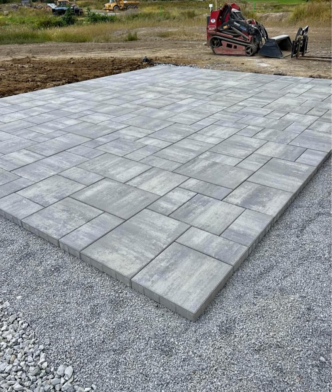 Gray pavers laid on gravel, with construction equipment in the background, likely a new patio or outdoor area.