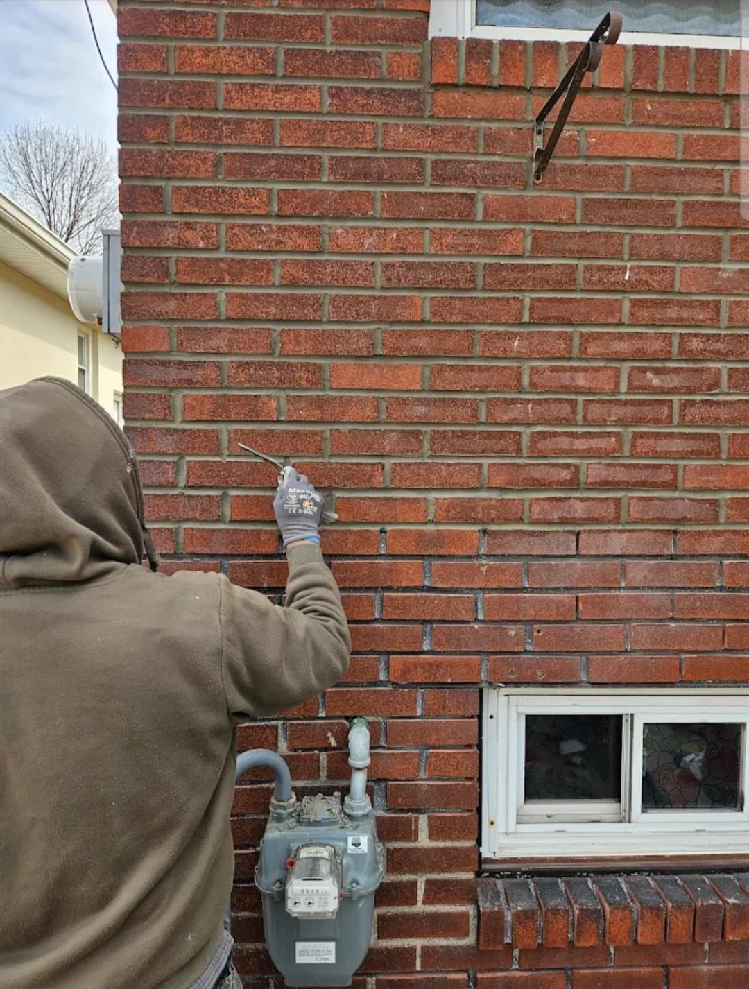 Person in hoodie pointing at a brick wall near a gas meter and window.