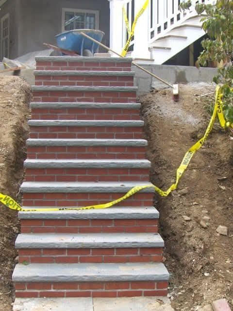 Brick staircase under construction with gray stone steps, surrounded by dirt and caution tape.