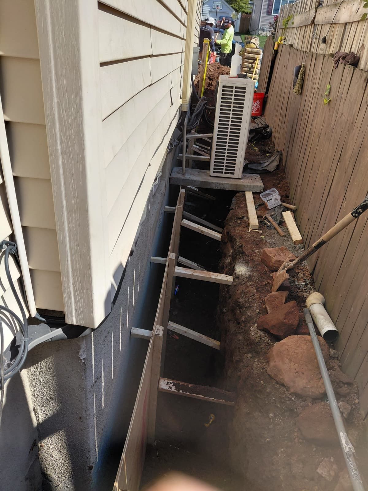 Construction site next to a house with a trench dug. Metal supports brace a foundation wall.