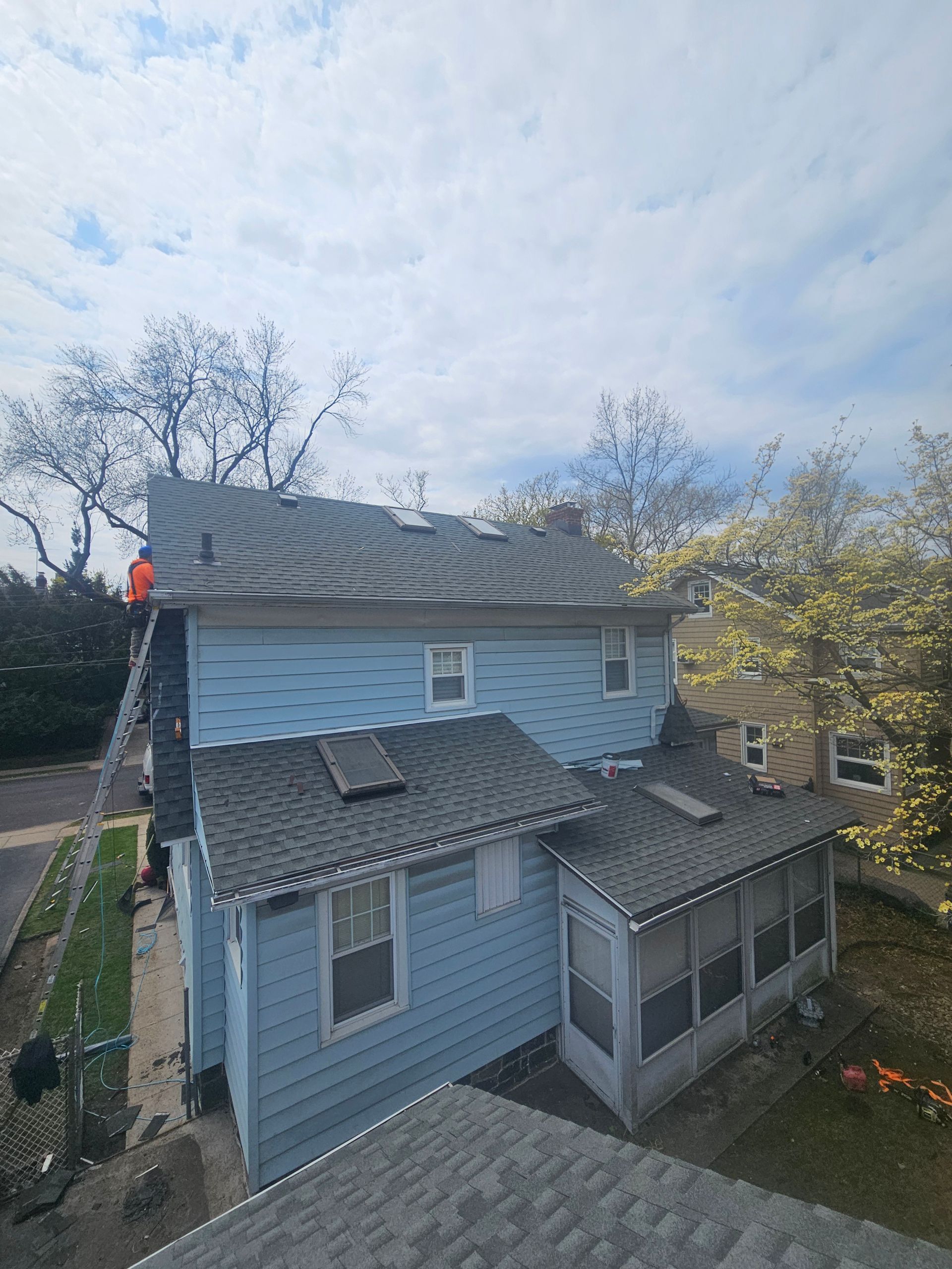 Blue house with dark gray roof, ladder, and person working on roof. Cloudy sky.