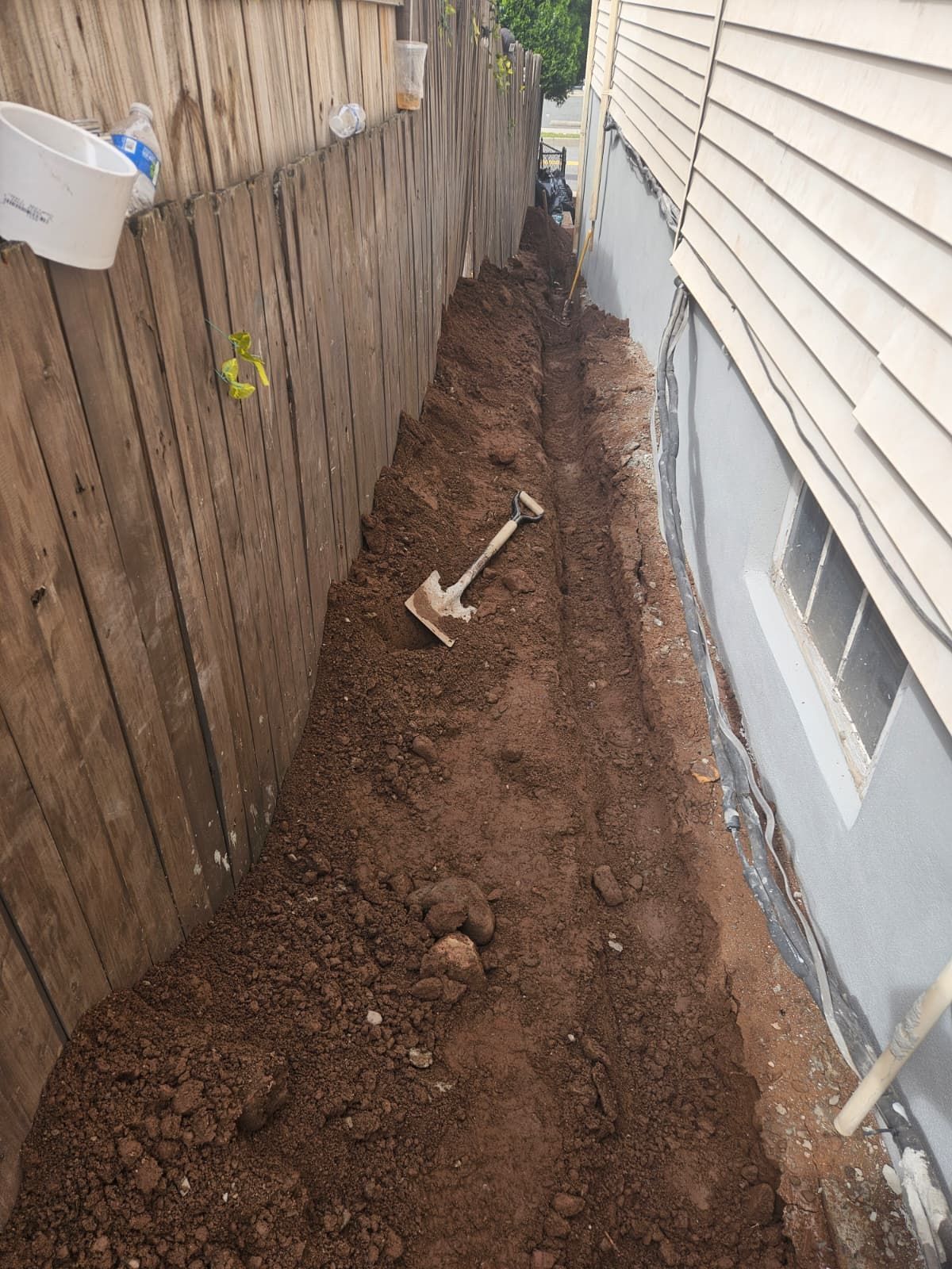 A narrow trench dug alongside a wooden fence and the side of a building, with a shovel resting in the dirt.