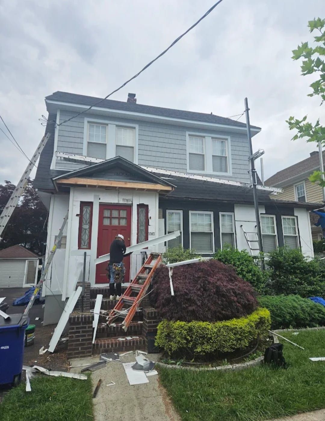 House with workers; roof repair. Man on steps, ladders, gray siding, white trim, red door. Overcast day.