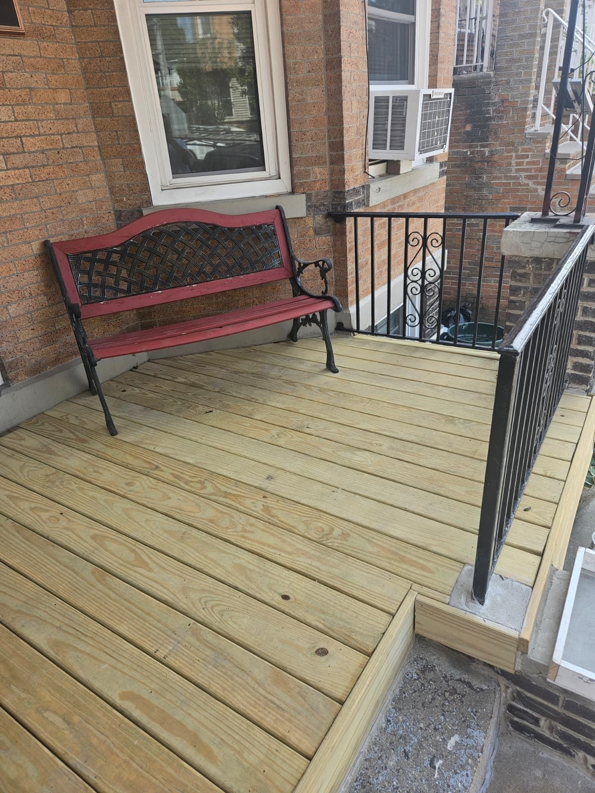 Wooden porch with a red bench and black railing against a brick building.