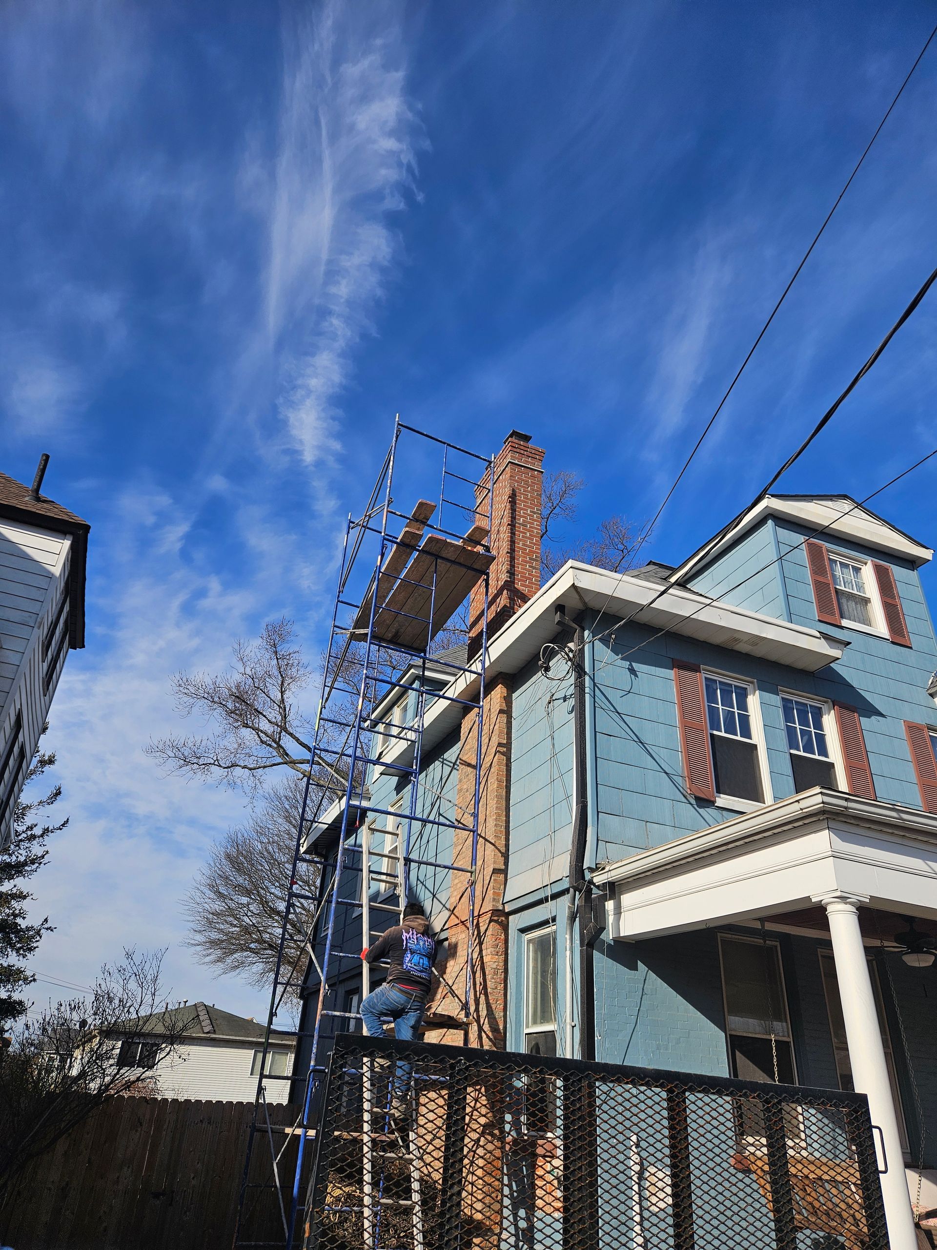 Person on scaffolding repairing a brick chimney on a blue house under a partly cloudy sky.