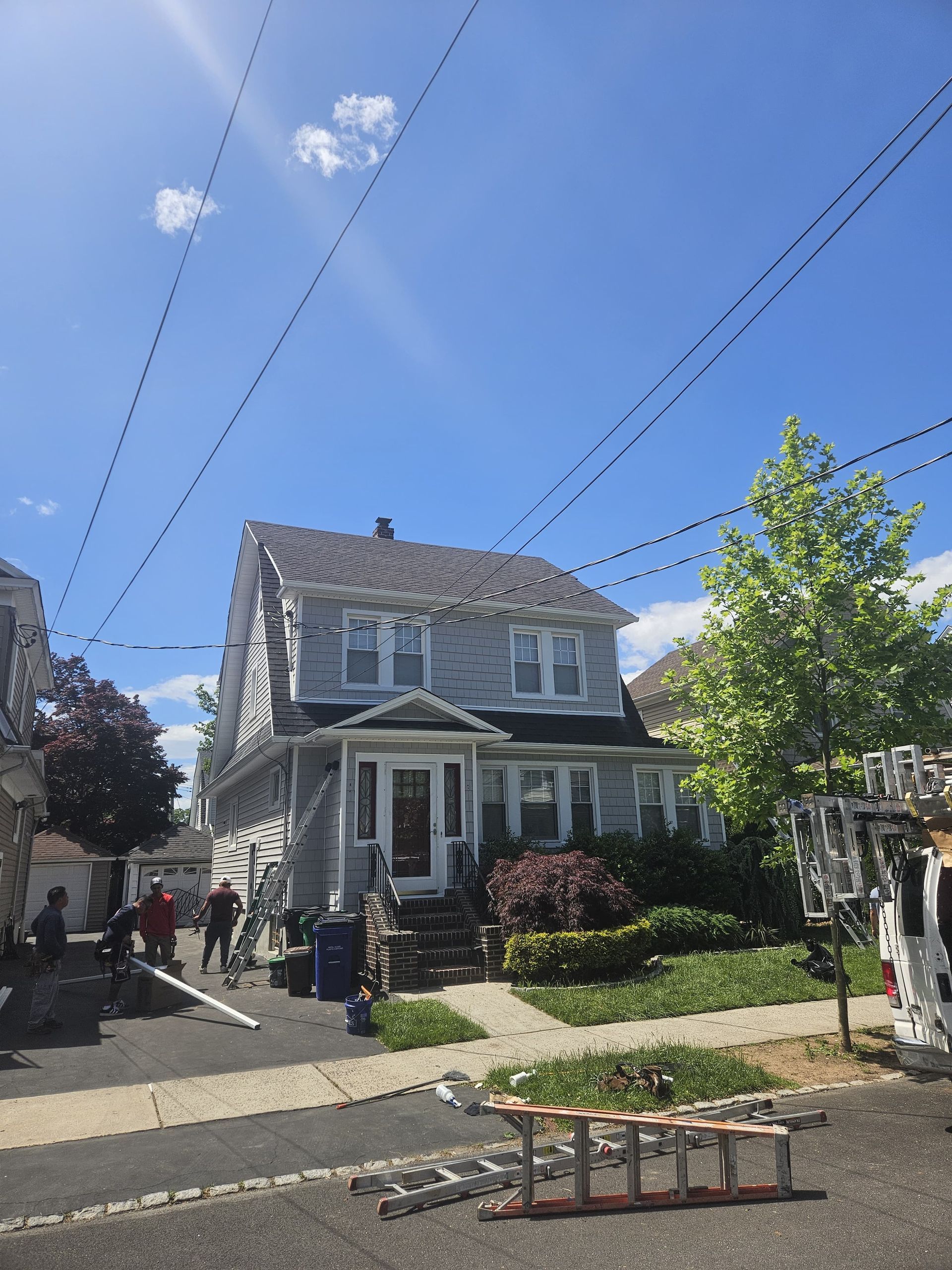 Two-story house with gray siding, blue sky, power lines. People and equipment on the street suggesting construction.