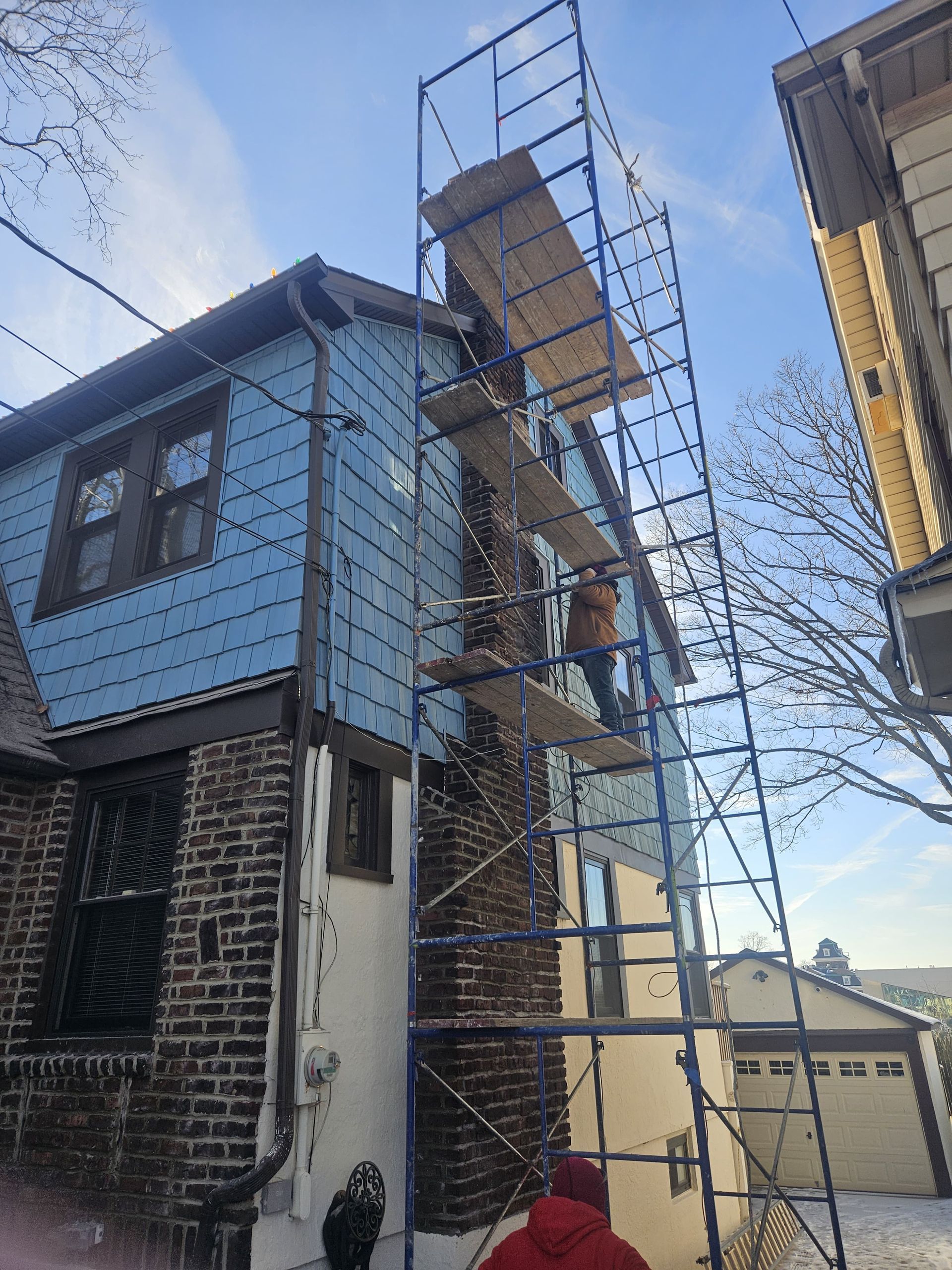 Scaffolding erected on a house, a person works on the upper platform. Blue siding and brick facade. Cloudy sky.