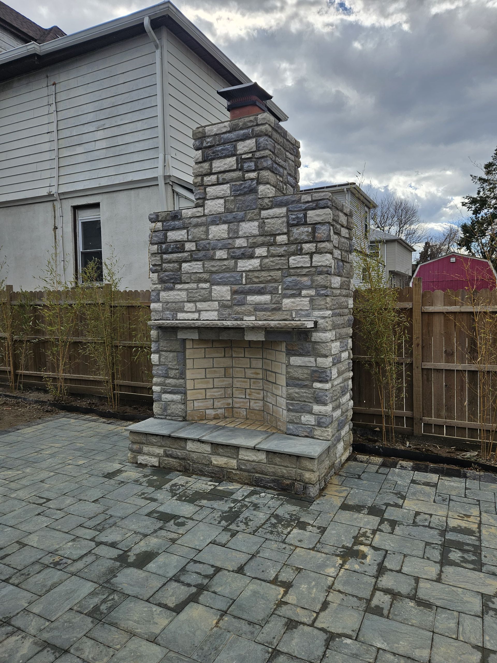 Stone outdoor fireplace on a paved patio, with a brick firebox and chimney, next to a wooden fence.