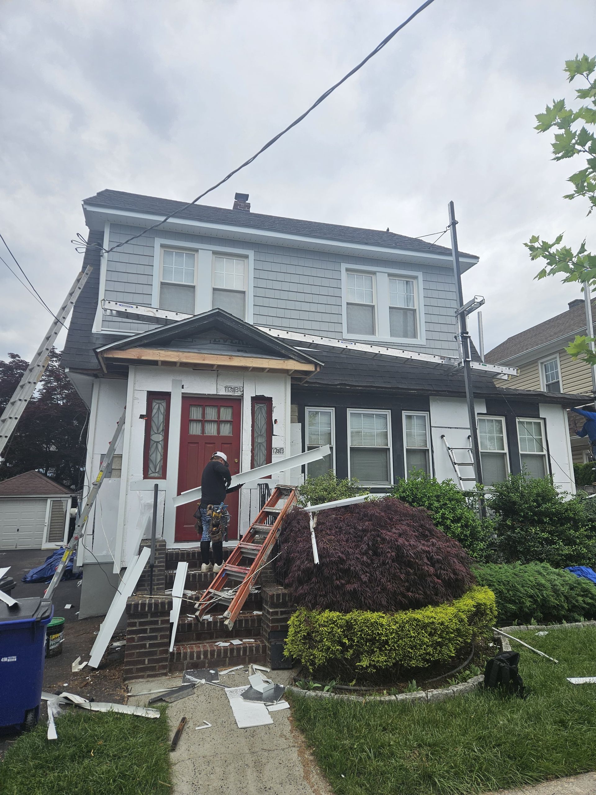 House exterior under construction, worker on ladder by red door; siding, roof, and trim being replaced.