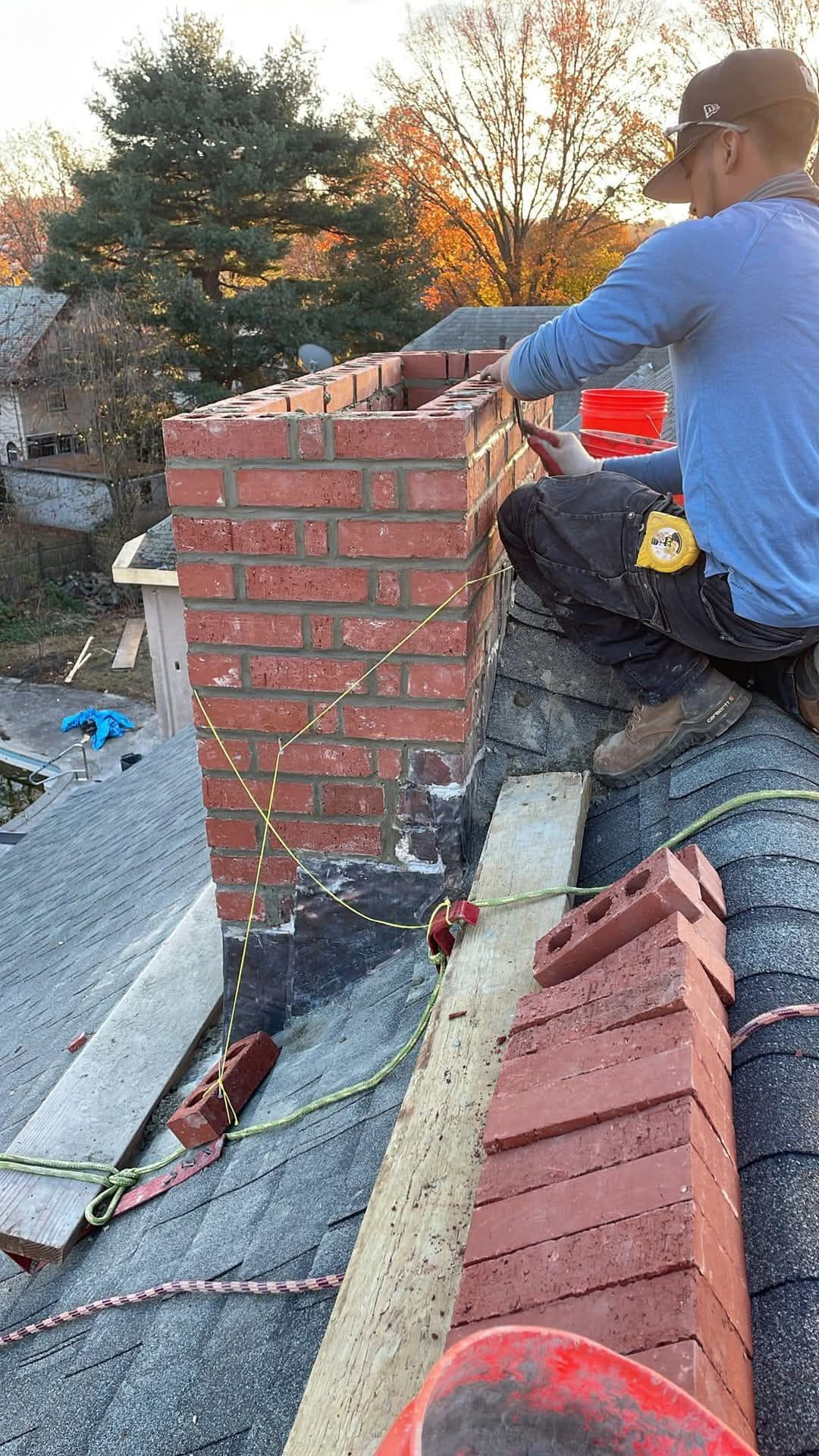 Person building a brick chimney on a rooftop. Bricks are red. The person is wearing a blue shirt.
