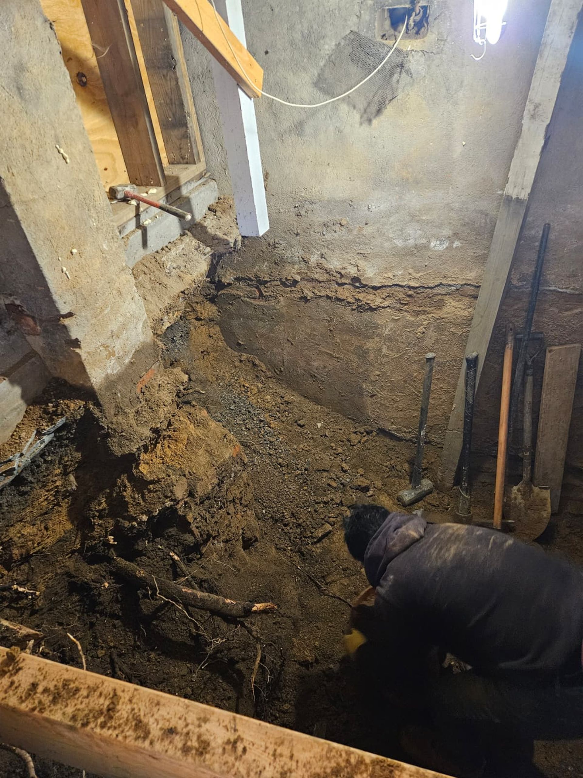Person working in a dirt excavation next to a concrete wall and wooden supports.
