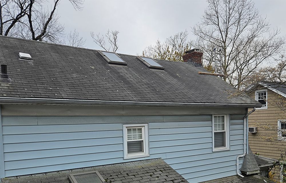 Blue house with a weathered roof, three skylights, and a brick chimney under an overcast sky.