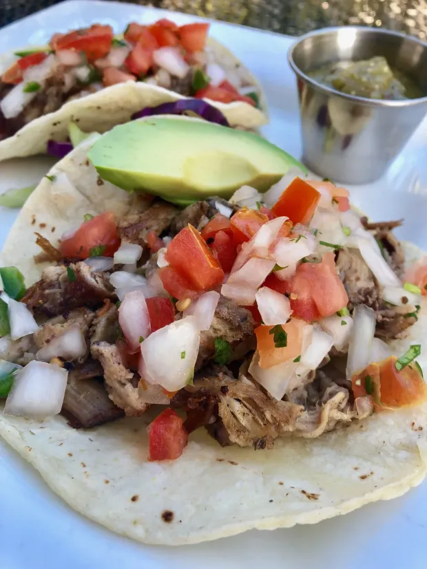 Tacos topped with shredded meat, diced tomatoes, onions, cilantro, and avocado slices, with lime on the side
