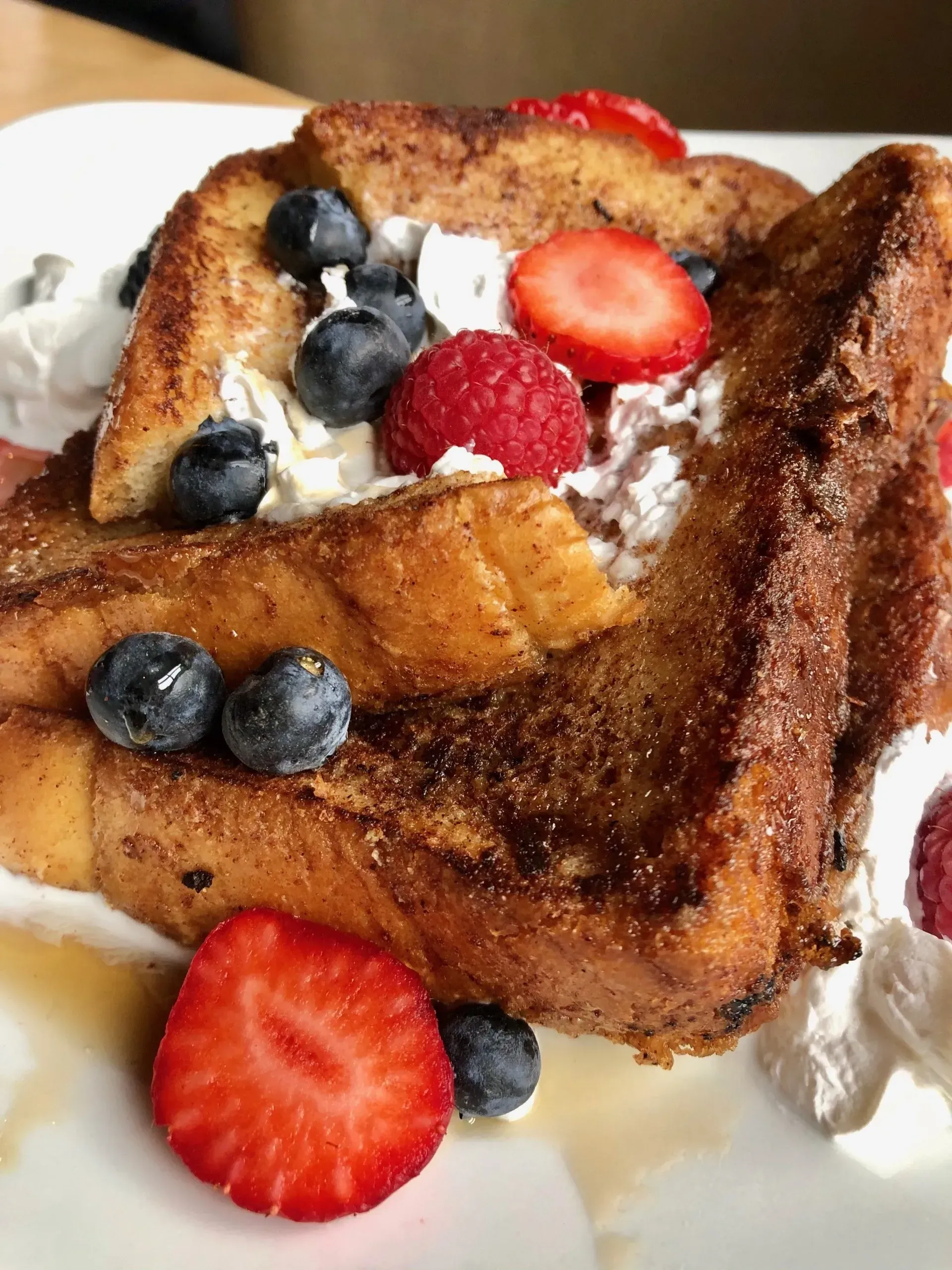 French toast topped with whipped cream, blueberries, and sliced strawberries, drizzled with syrup