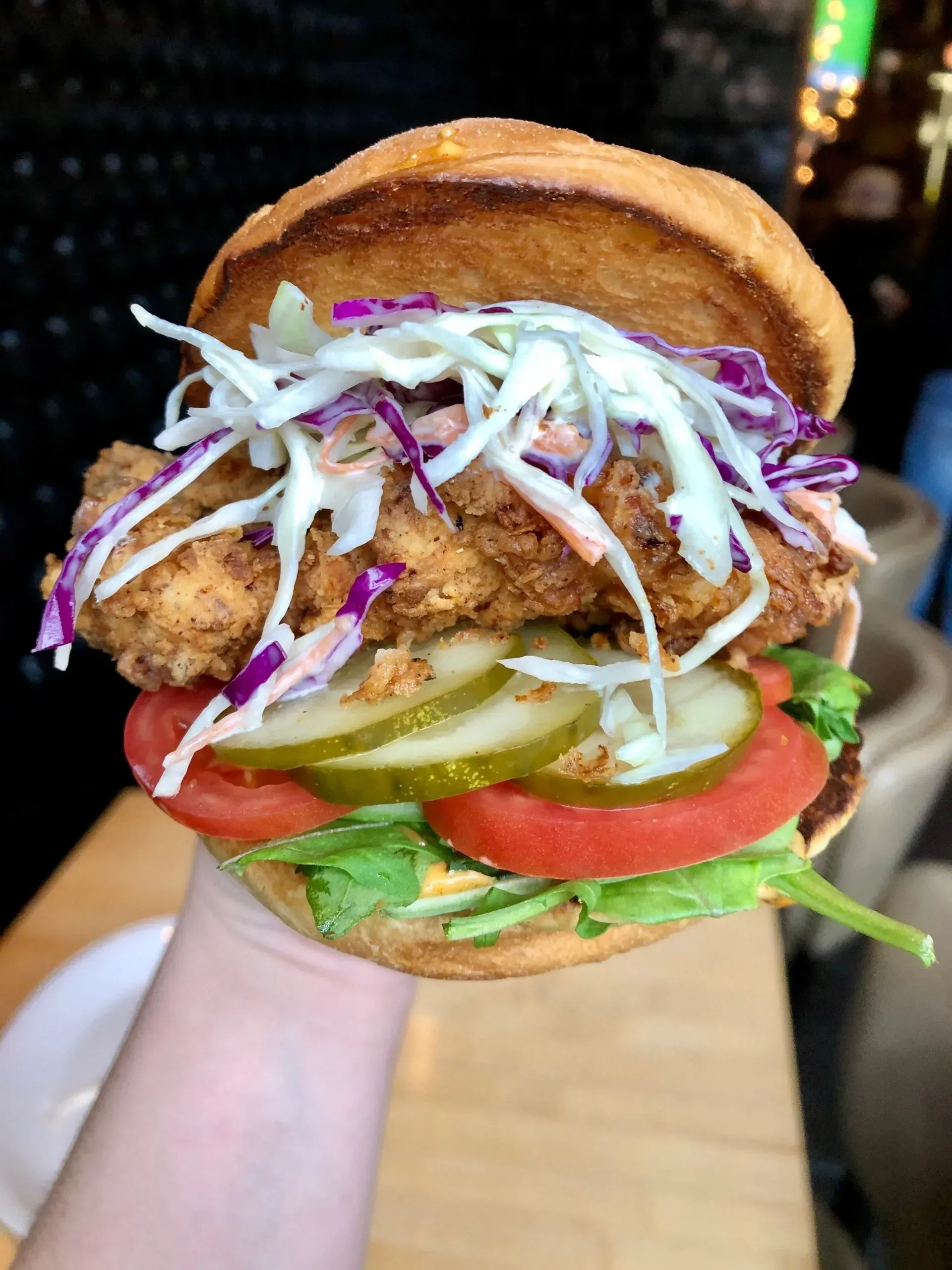 A hand holding a stacked burger with lettuce, tomato, pickles, coleslaw, and a toasted bun.