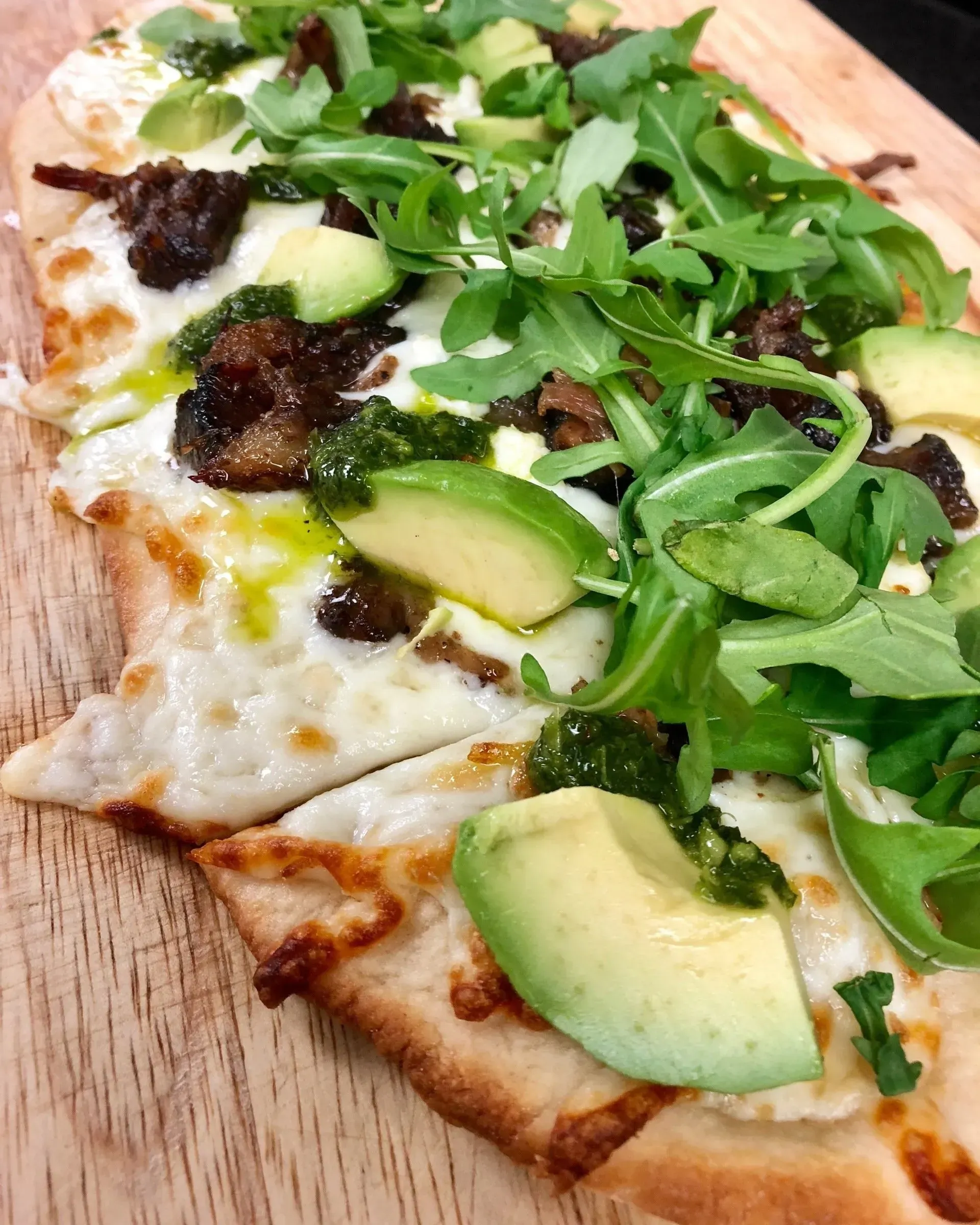 Slice of pizza topped with arugula, avocado, and melted cheese on a wooden board