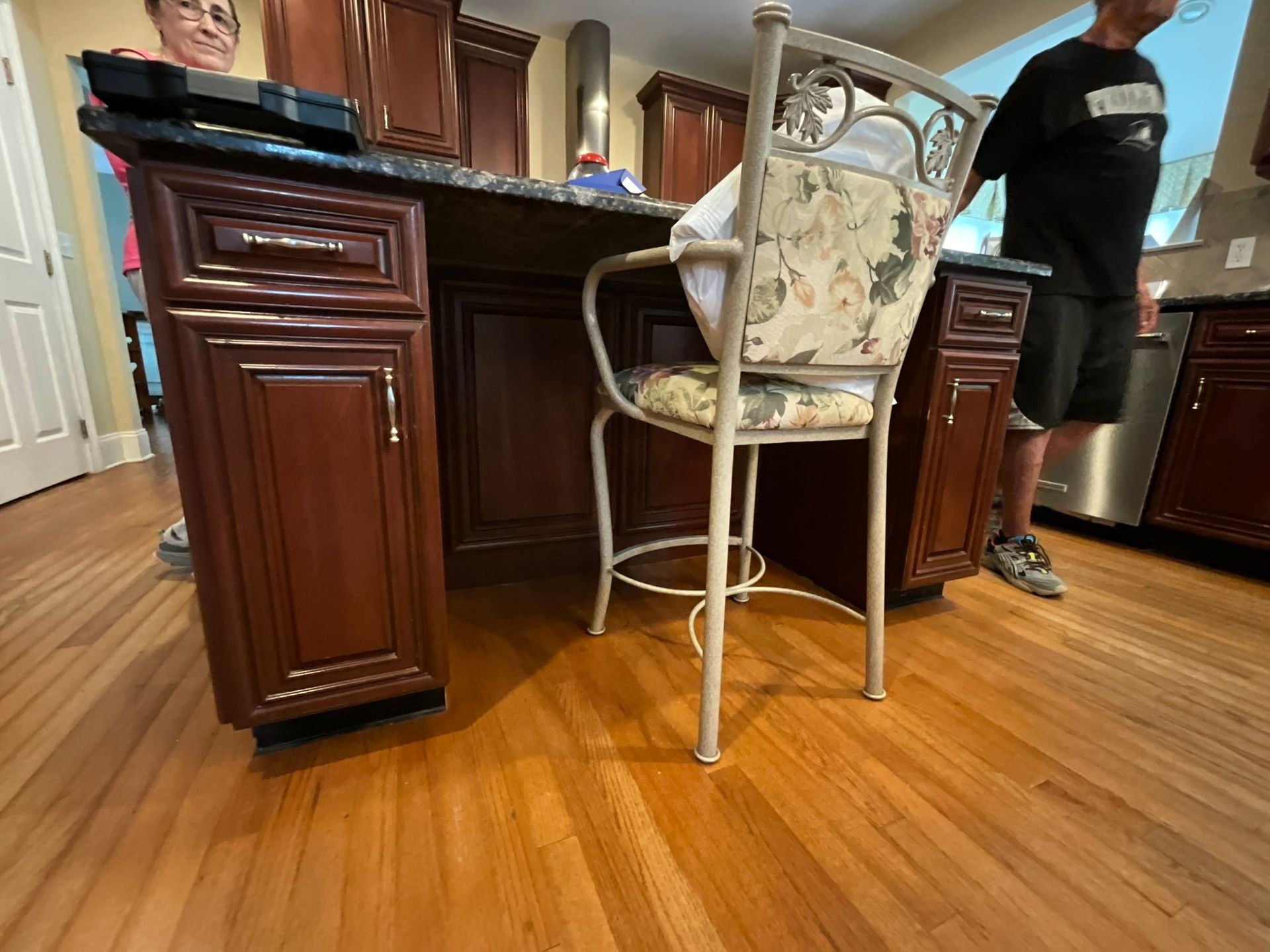 Kitchen island with dark wood cabinets, a granite countertop, and a floral patterned chair. Two people stand nearby.