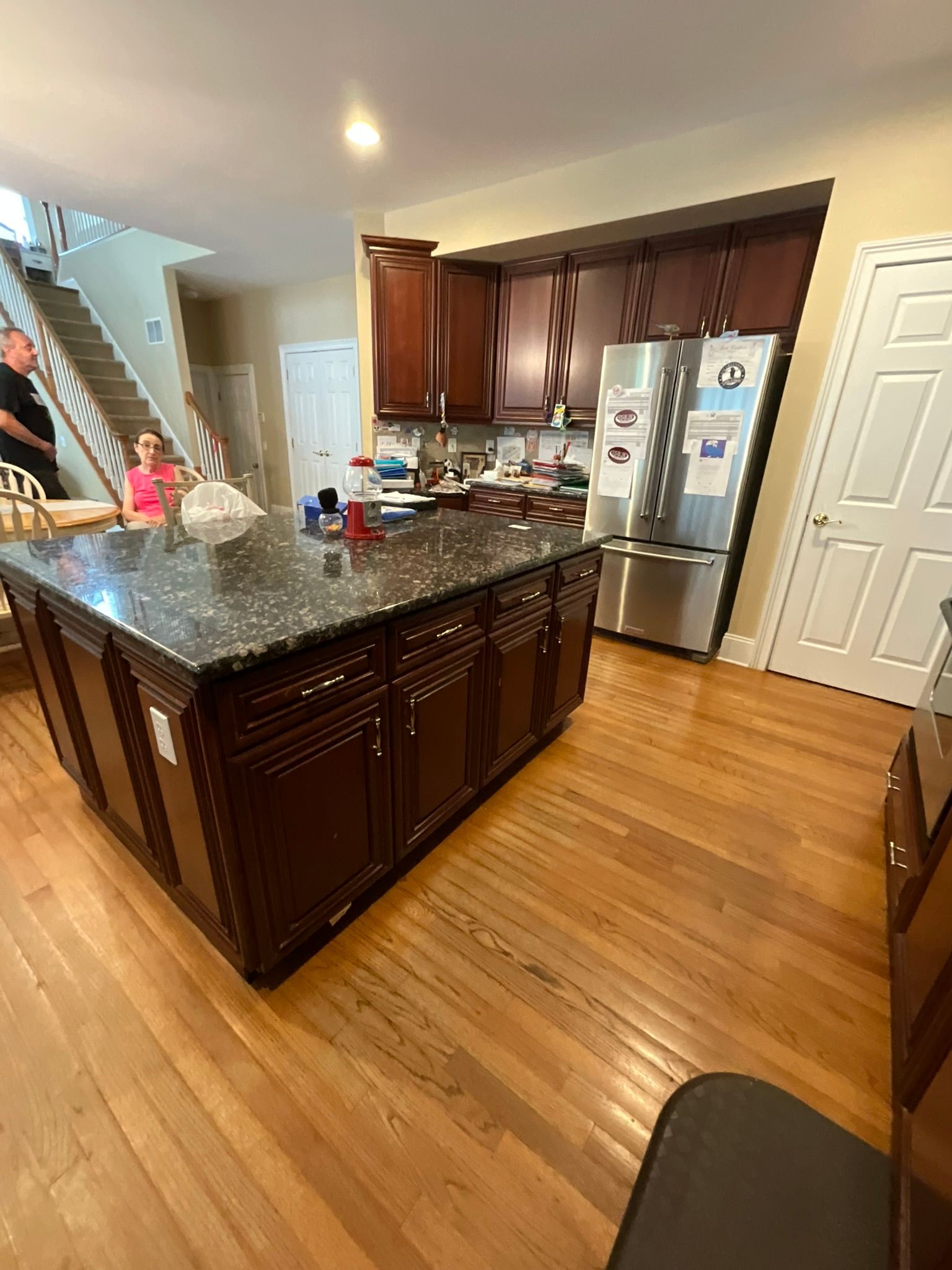 Kitchen with dark wood cabinets, island, granite countertop, stainless steel fridge, hardwood floors, and two people.