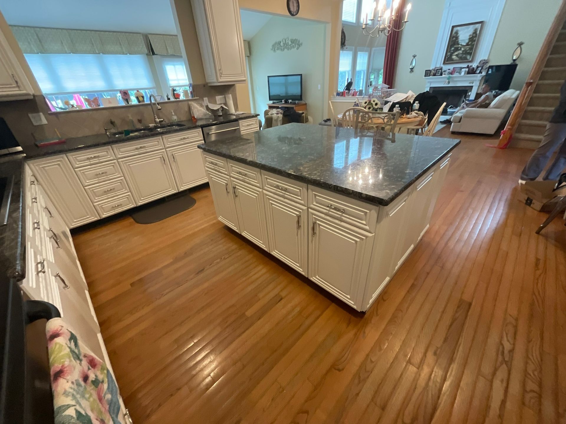 Spacious kitchen with white cabinets, a dark granite island, and hardwood floors. Sunlight streams in from windows.