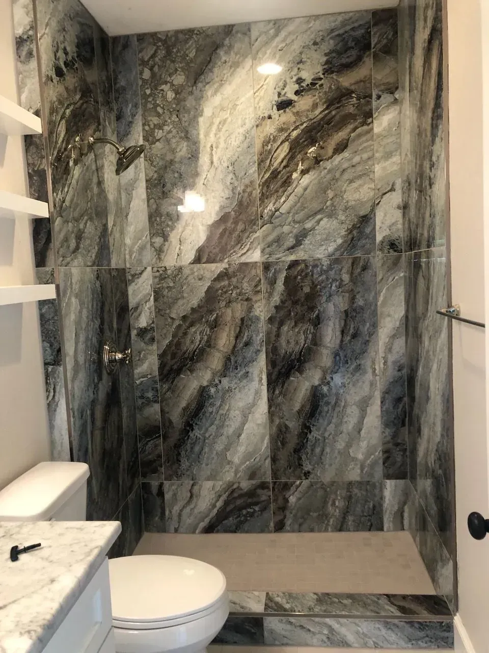 Bathroom with patterned marble shower tile, white toilet, and shelves.