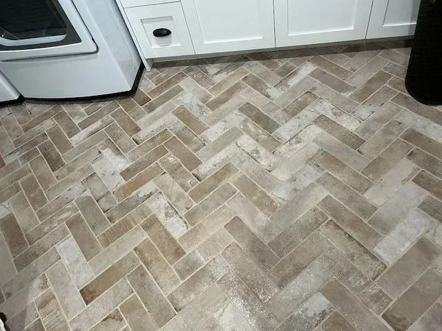 Herringbone patterned tile floor in a laundry room, with white cabinets and a washing machine visible.