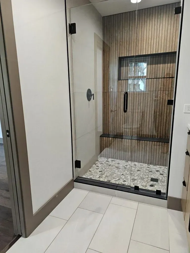Modern bathroom with glass shower enclosure, wood-look accent wall, pebble floor, and white tile.