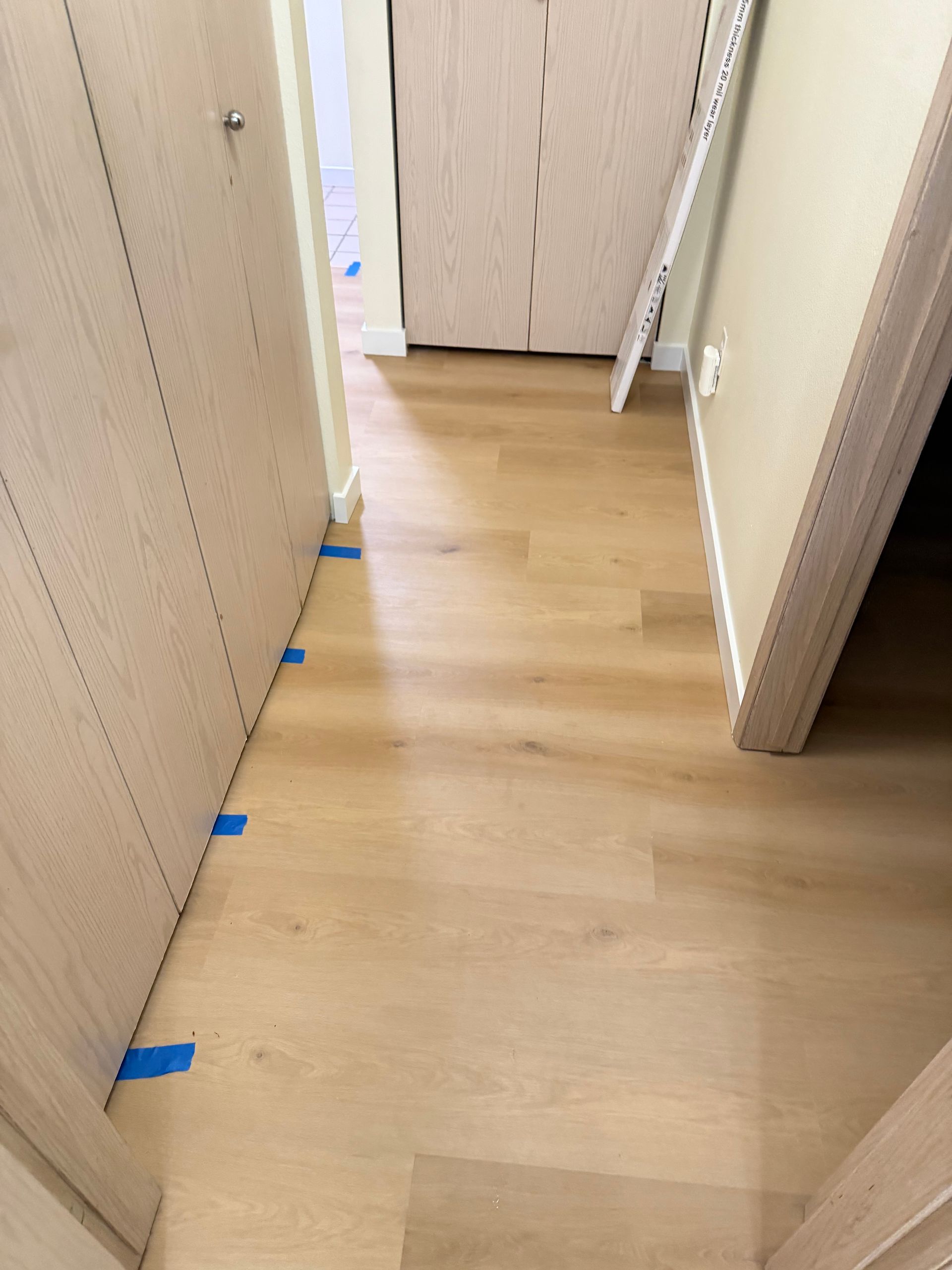 Hallway with light wood-look flooring and beige walls; doors on either side, blue tape along baseboard.