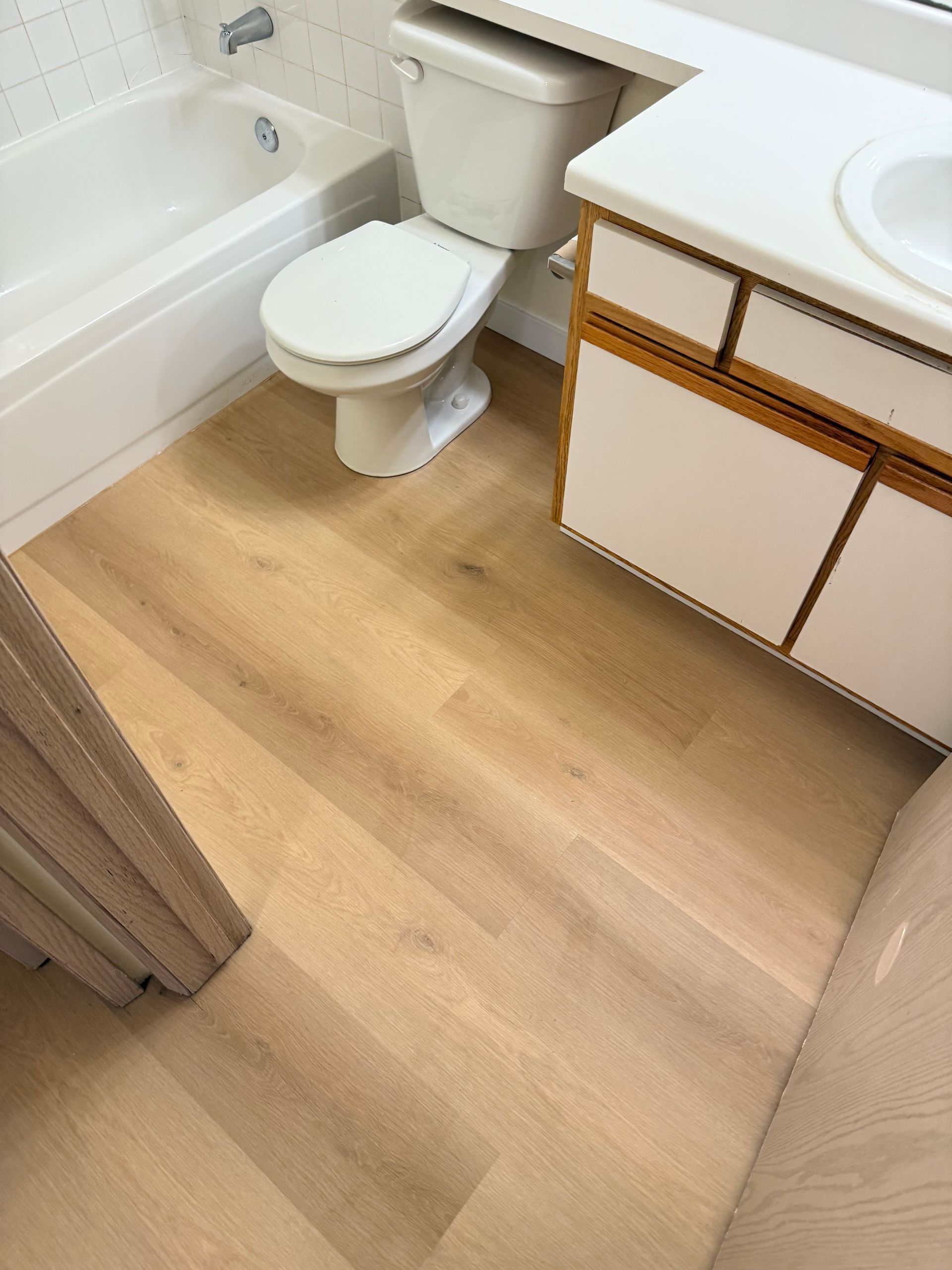 Bathroom with light wood-look flooring, white tub, toilet, and vanity with a light countertop.