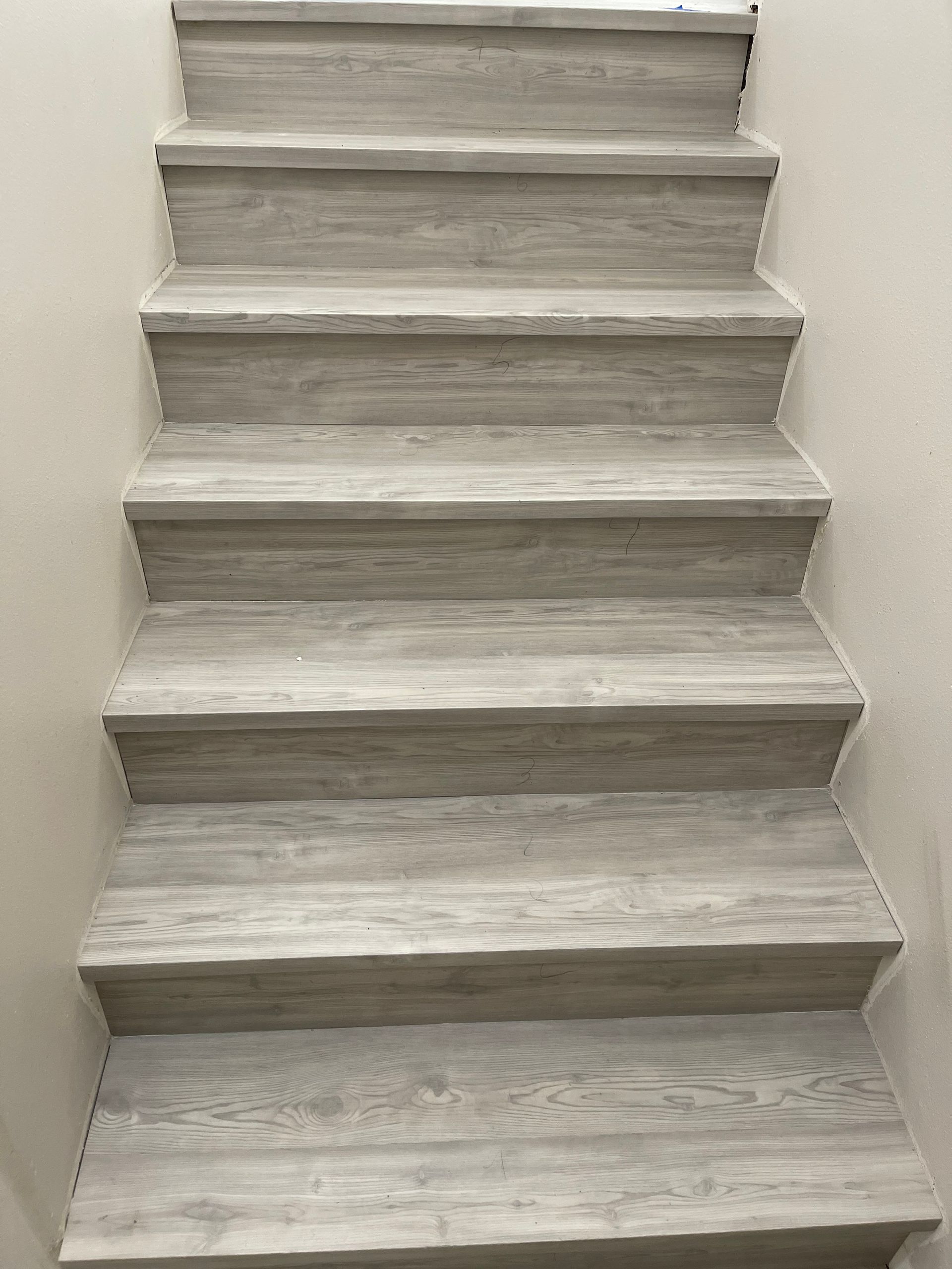 Staircase with light gray, wooden-look treads and risers, ascends between off-white walls.