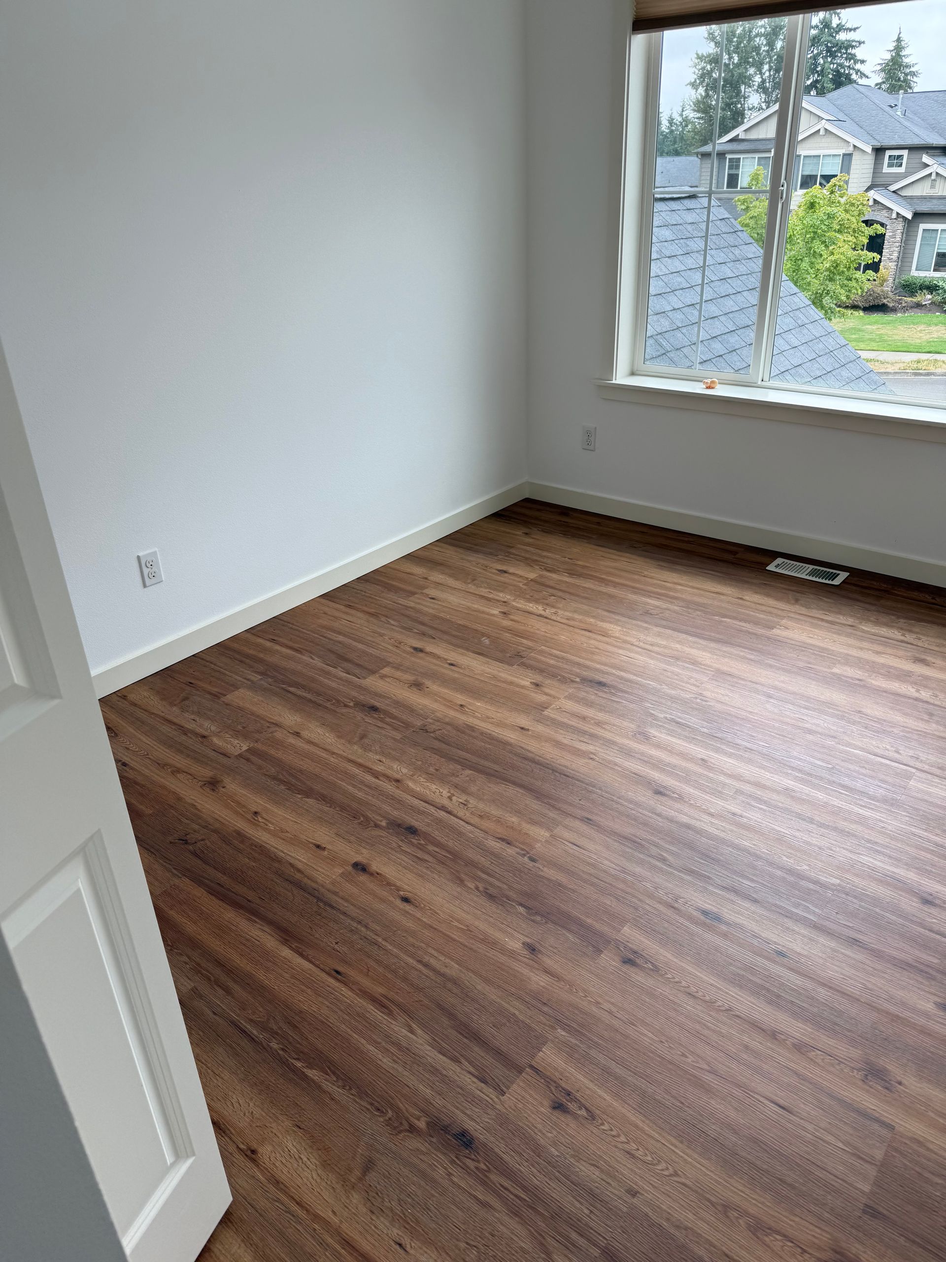 Empty room with wood-look flooring and a window overlooking houses. White walls and door.