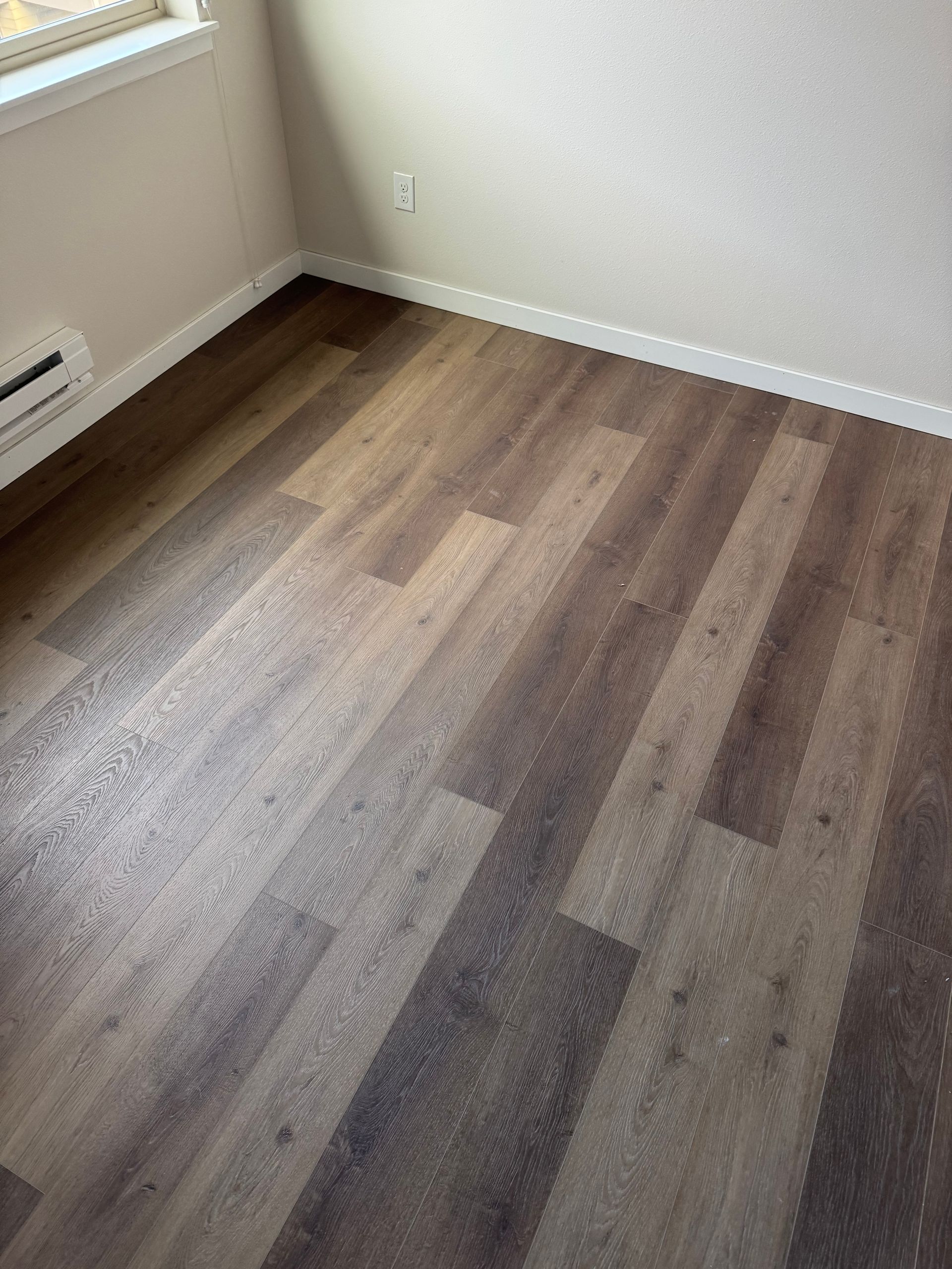 Empty room with dark brown wood-look flooring, white baseboards, and pale walls. A window is in the top left corner.